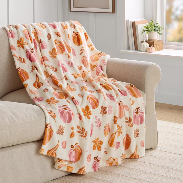 A cream-colored throw blanket with orange and pink pumpkin and leaf patterns draped over a beige sofa in a bright living room with a window and potted plant.