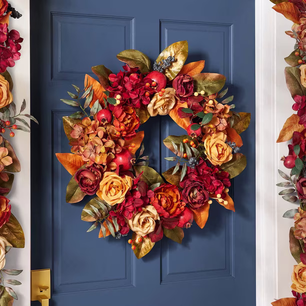 A fall wreath with red, orange, and yellow flowers, green leaves, and small berries hangs on a dark blue door with a gold handle.