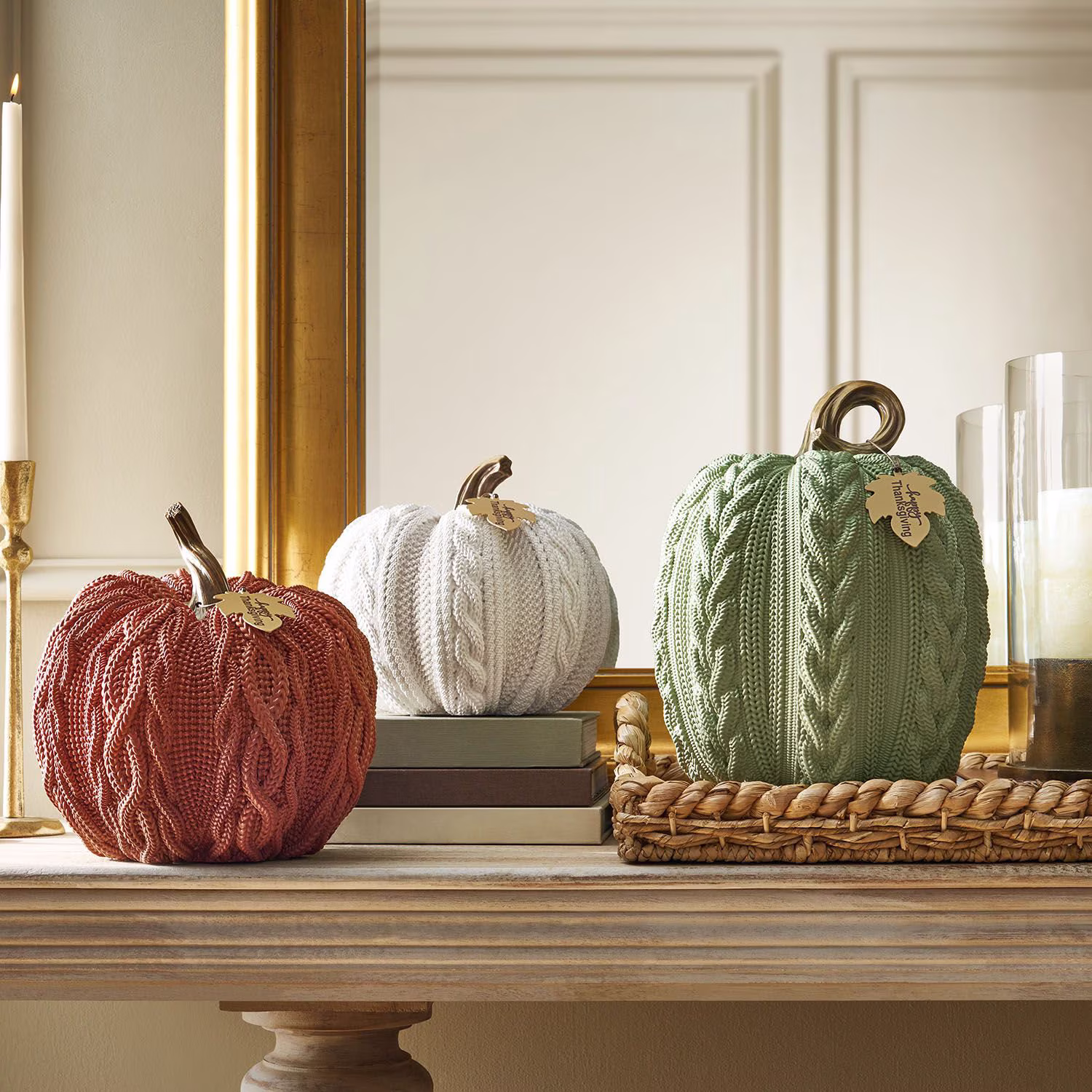 Three knitted decorative pumpkins in orange, white, and green sit on a mantel with a candle, books, and tray in a cozy, elegant room. Each pumpkin has a gold stem and a small gold leaf tag.