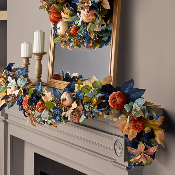 A decorated mantel with autumn-themed garland featuring orange pumpkins, blue and yellow foliage, berries, and candles, with a matching wreath reflected in a framed mirror above.