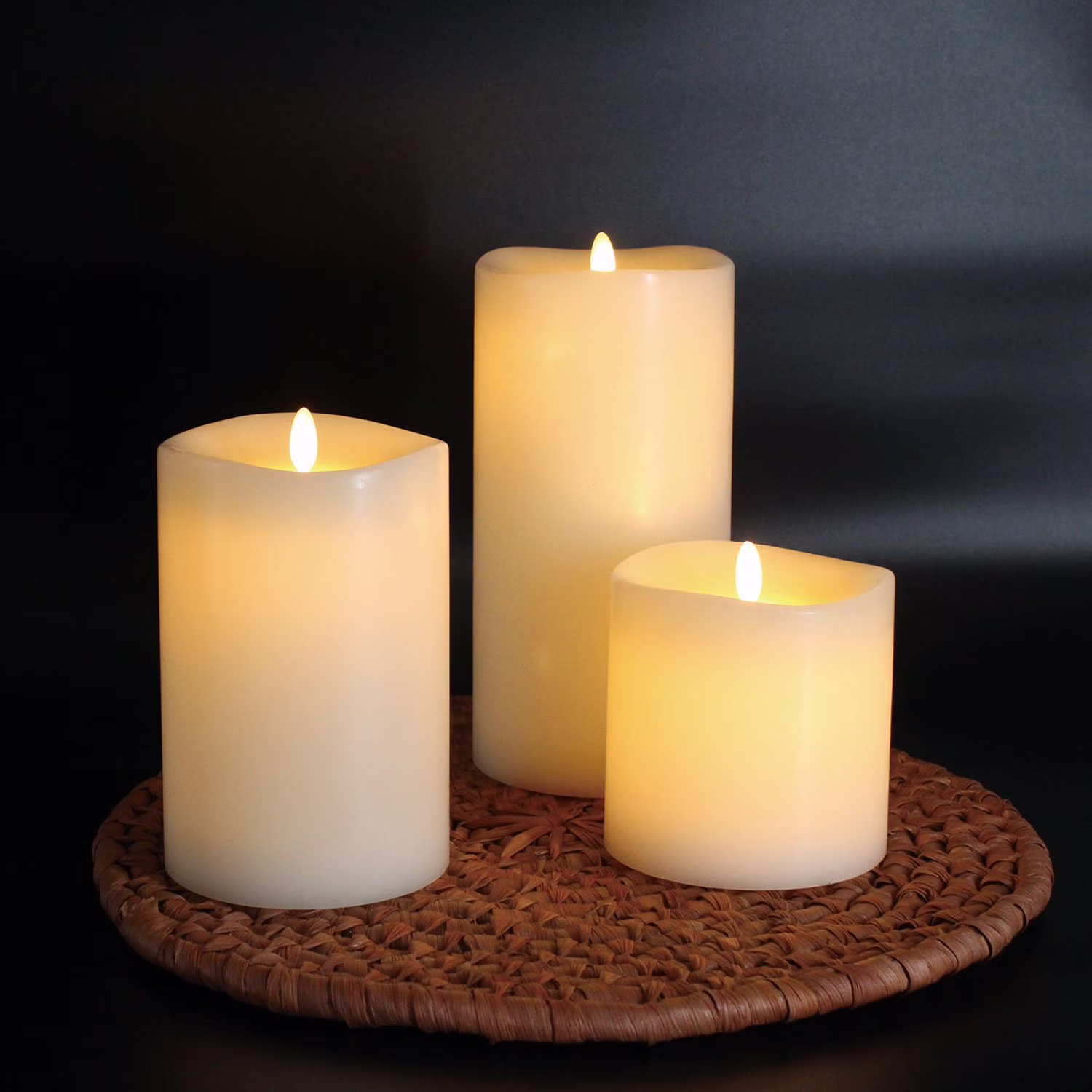 Three flameless pillar candles of varying heights, glowing with warm light, are placed on a round woven mat against a dark background.