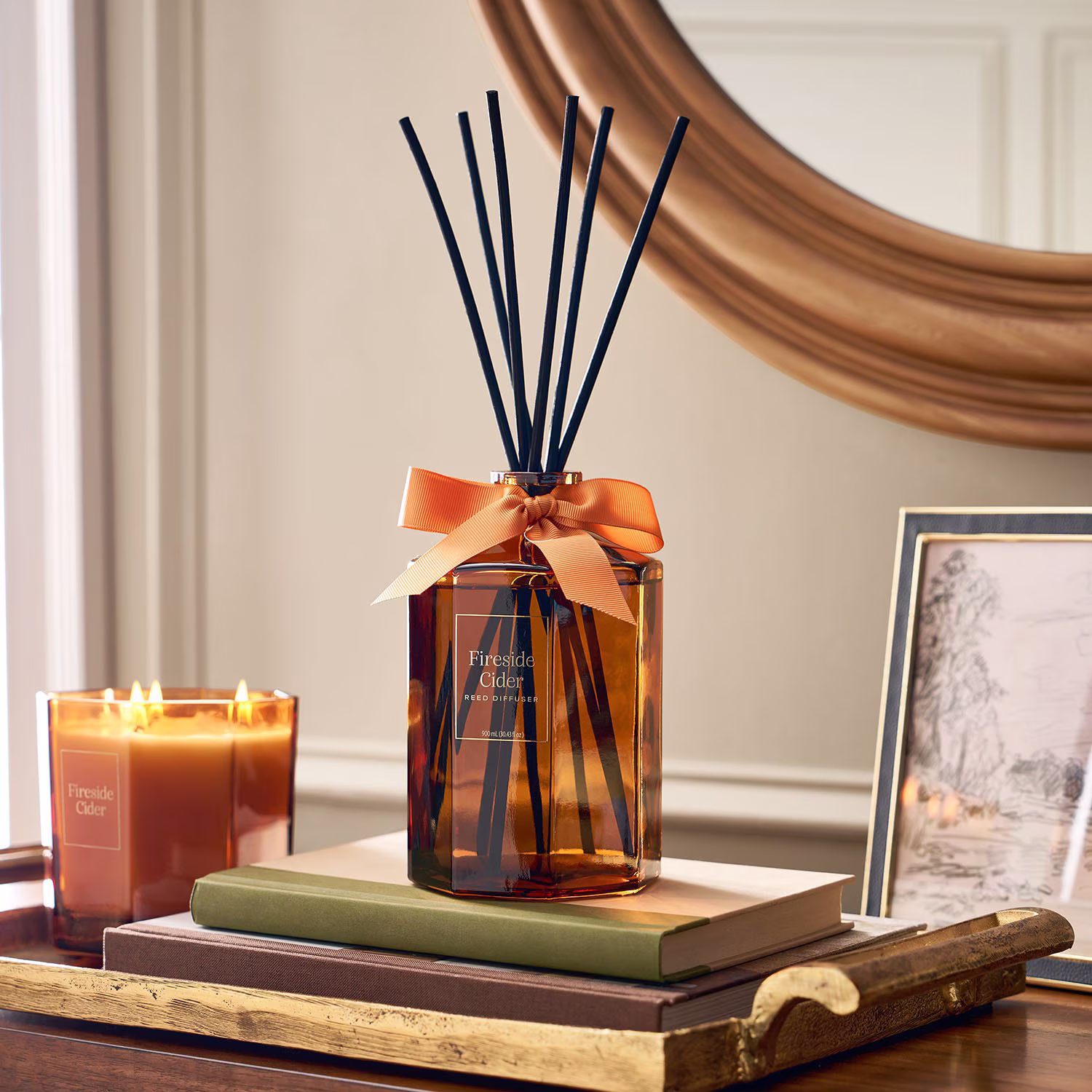 Amber reed diffuser with orange ribbon sits on stacked books beside a burning candle, on a gold tray. A framed picture and a round mirror are in the background, creating a cozy, elegant setting.