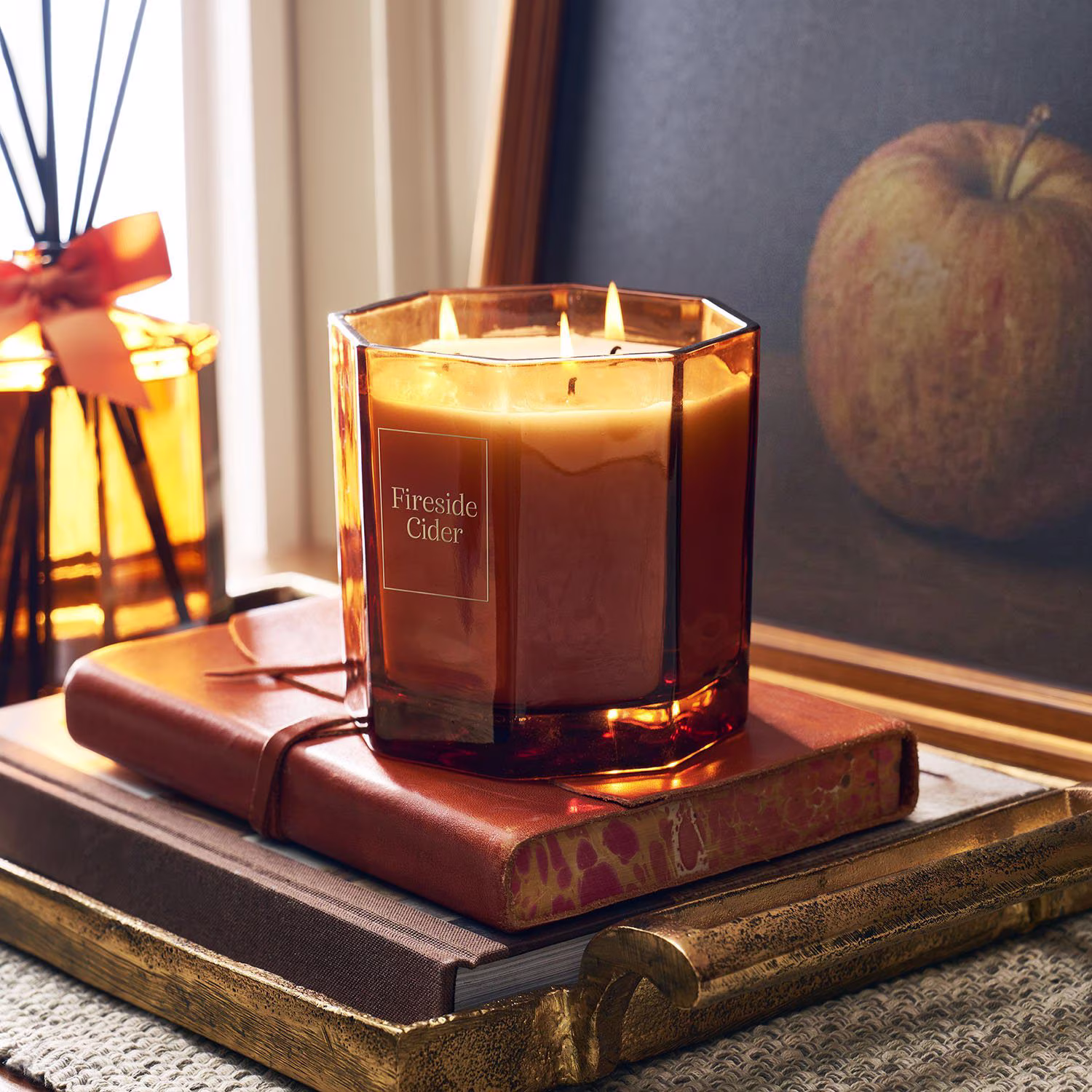 A lit “Fireside Cider” candle in a brown glass jar sits on stacked books next to a framed apple painting and a reed diffuser tied with an orange ribbon.
