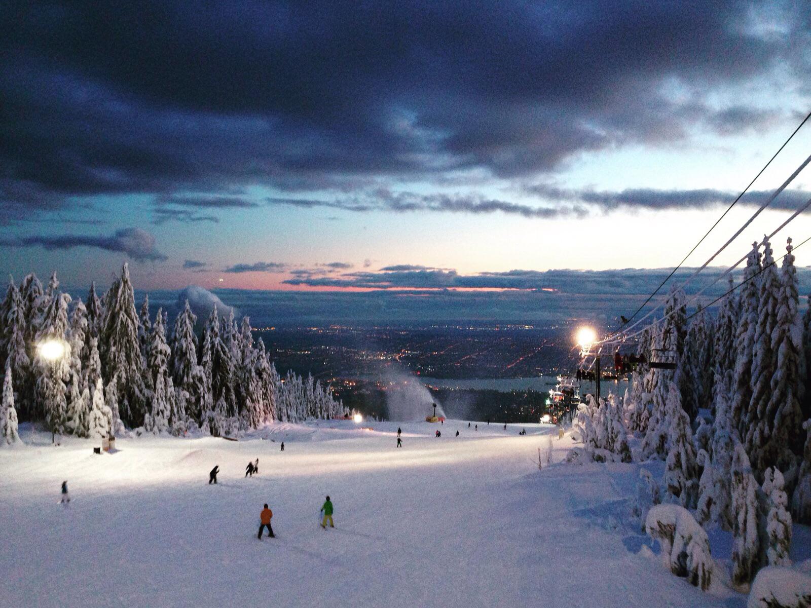Skiers descend a snowy slope lined with frosted trees at dusk, as city lights glow in the distance and a ski lift ascends on the right under a dramatic, cloud-filled sky.