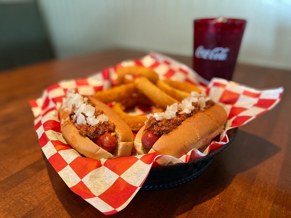 A basket with two chili dogs topped with chopped onions, several onion rings, and a red checkered paper liner. A red Coca-Cola cup sits in the background on a wooden table.
