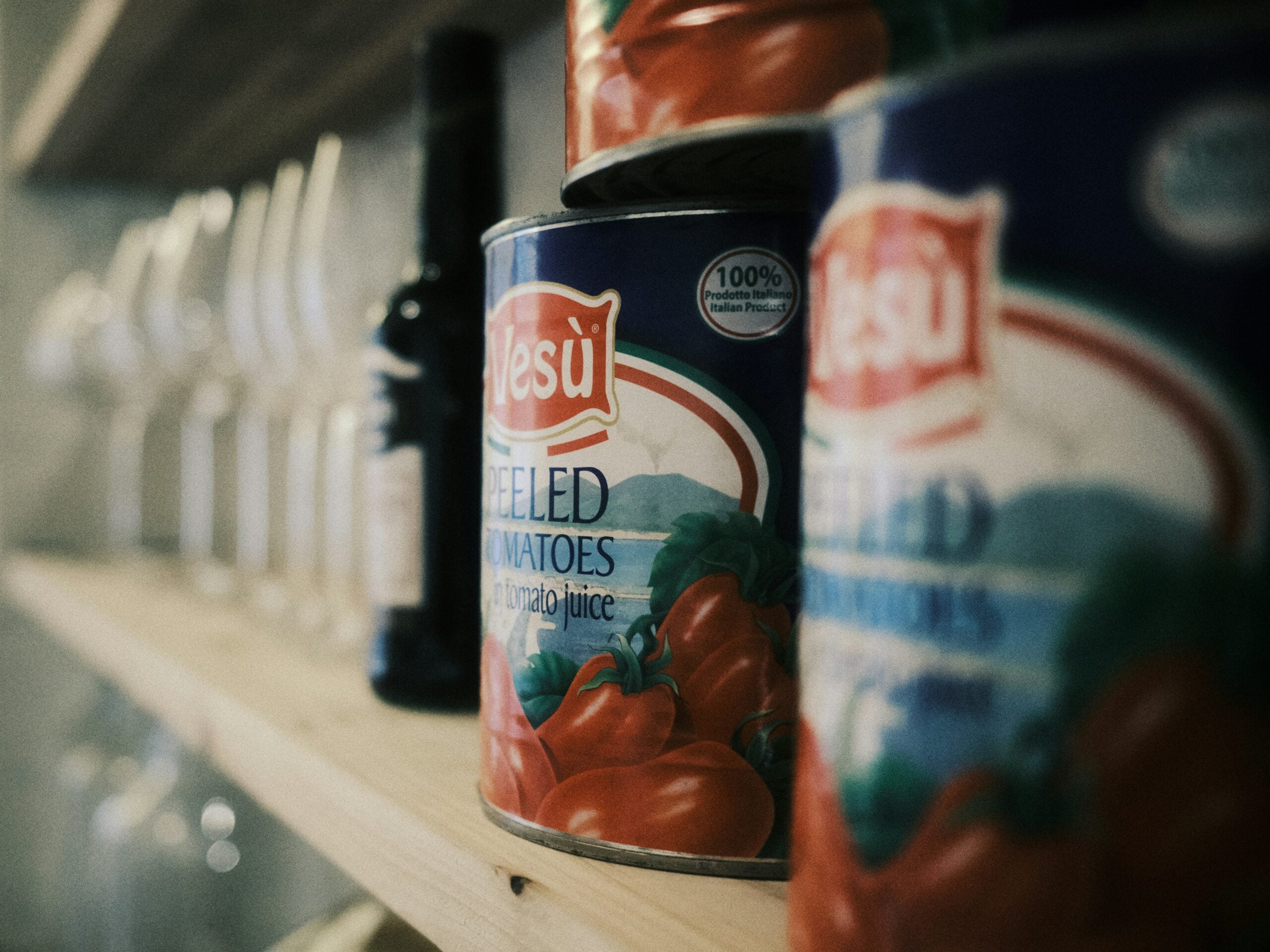 Close-up of cans of peeled tomatoes on a wooden shelf, with bottles and glassware blurred in the background. The label on the can reads “Yesu Peeled Tomatoes in Tomato Juice.”