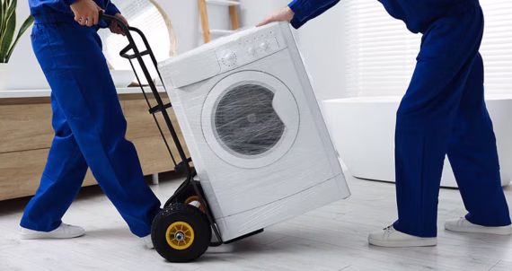 Two people in blue uniforms use a hand truck to move a washing machine wrapped in plastic across a light-colored floor in a bright room, highlighting some of the convenient services available with free Costco membership perks.