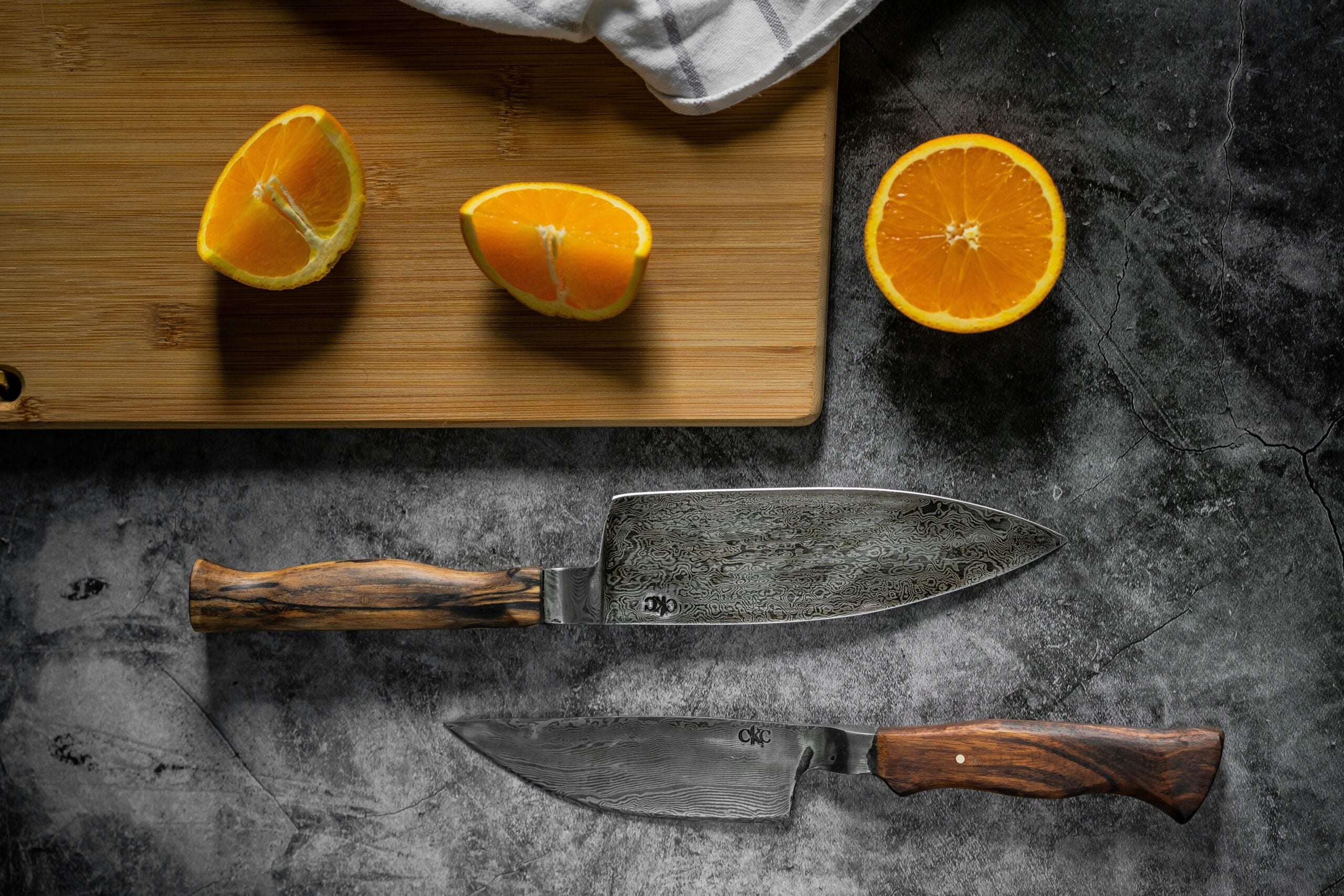 Two Damascus steel knives with wooden handles are placed on a gray stone surface next to a wooden cutting board and three orange halves, one on the board and two on the stone. A white cloth is partially visible at the top of the image.