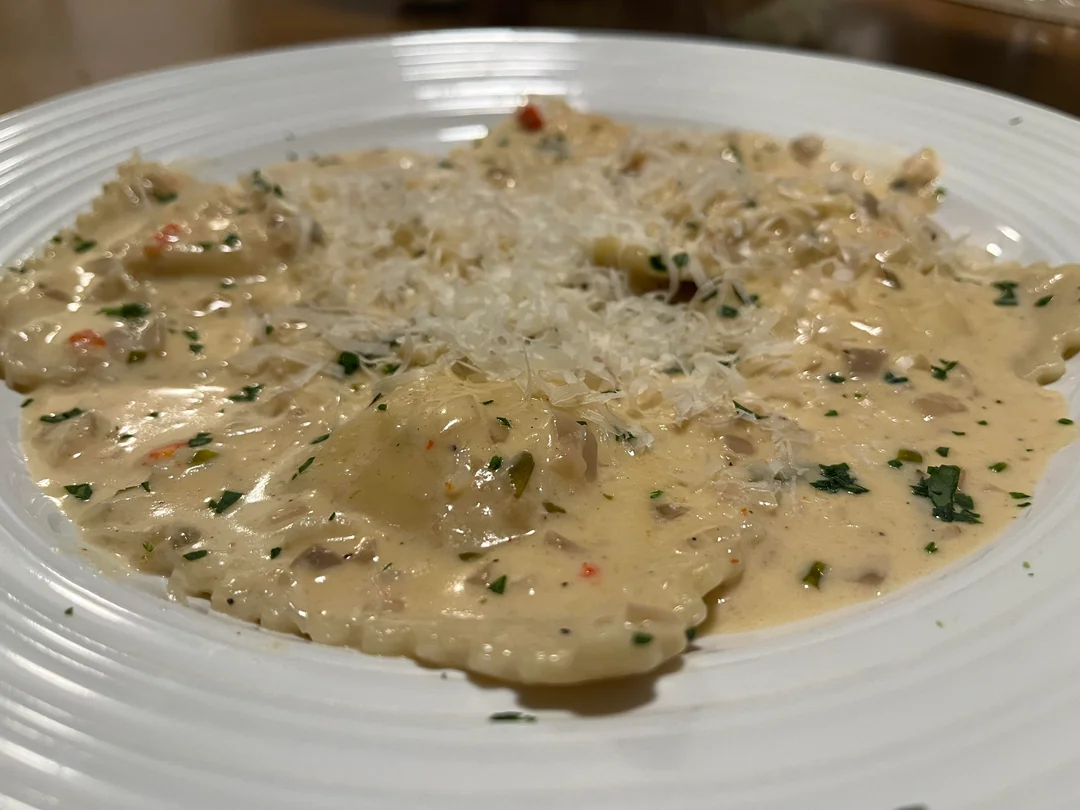 A close-up of ravioli in a creamy white sauce, topped with grated cheese and sprinkled with chopped herbs, served on a white plate.
