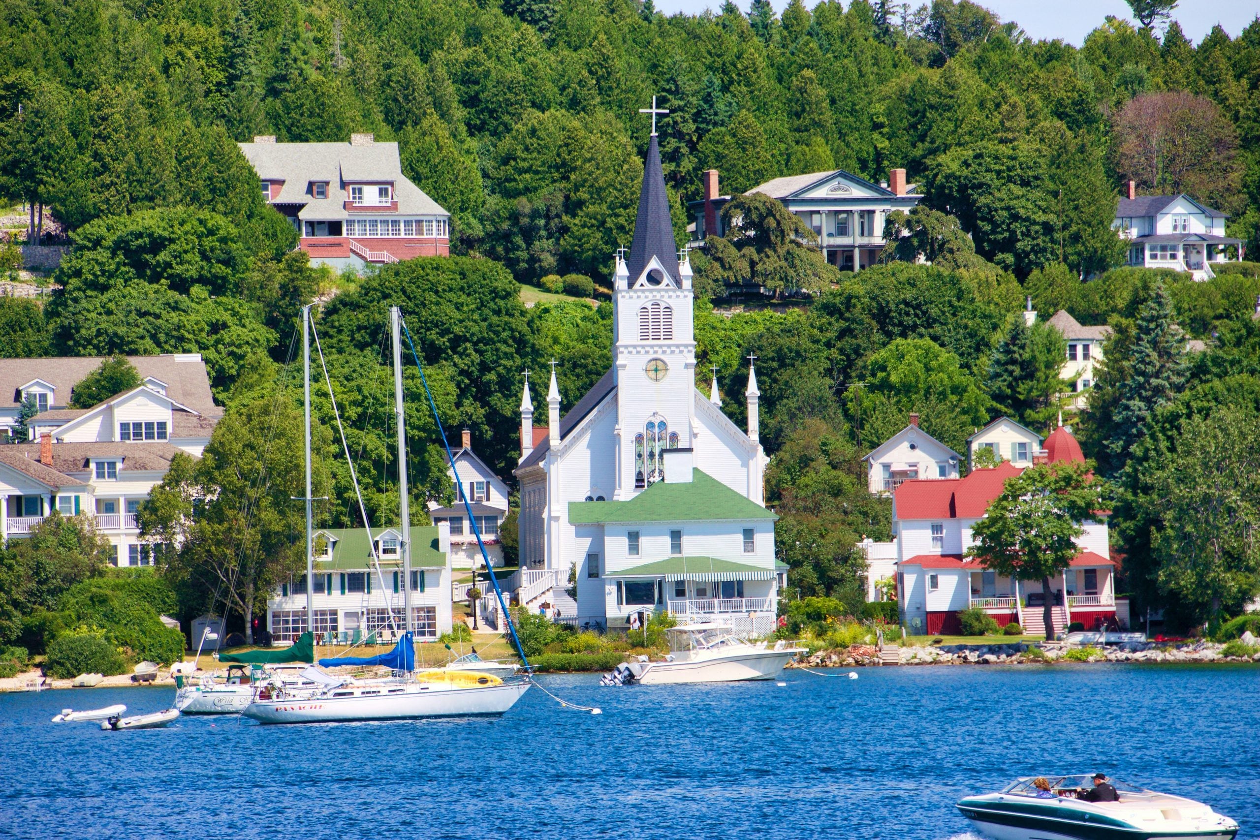 White church with a tall steeple stands among houses and lush green trees by a blue waterfront. Several boats are anchored in the water in the foreground.
