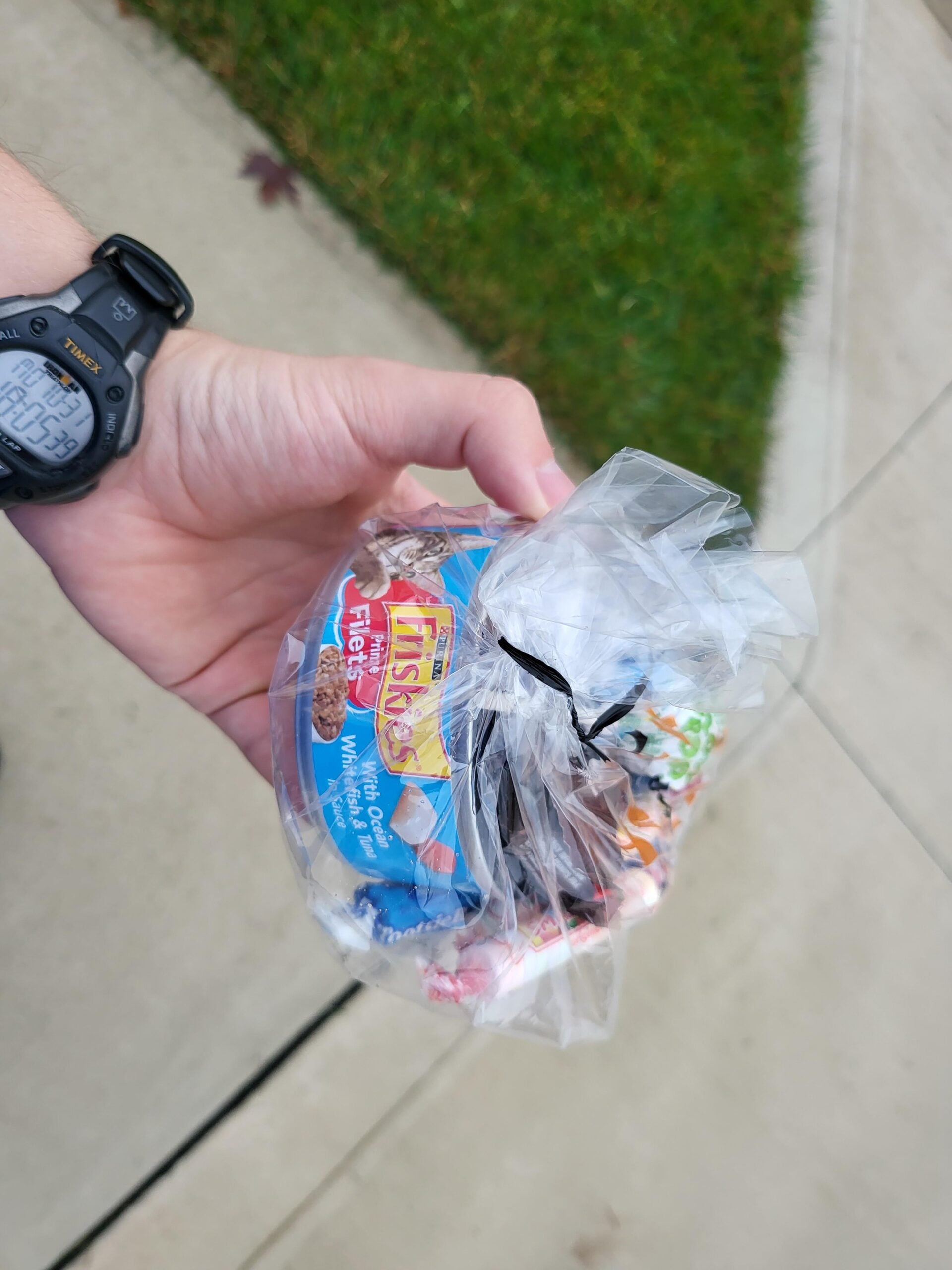 A hand holds a small clear plastic bag tied with a black twist tie, filled with assorted candies, including Tootsie Rolls. The background shows a sidewalk and grass.