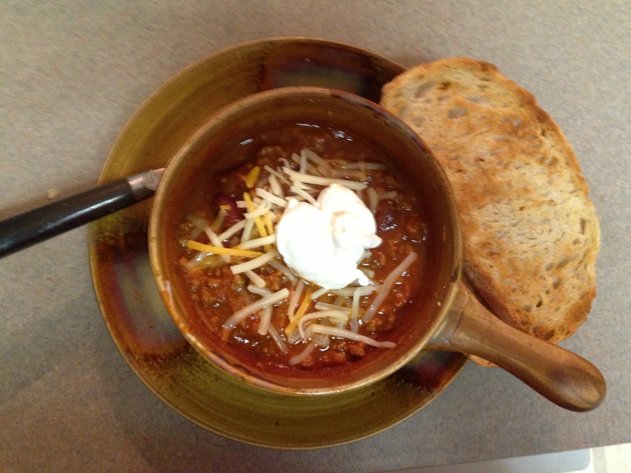A bowl of chili topped with shredded cheese and sour cream, served with a slice of toasted bread on the side, placed on a brown plate.