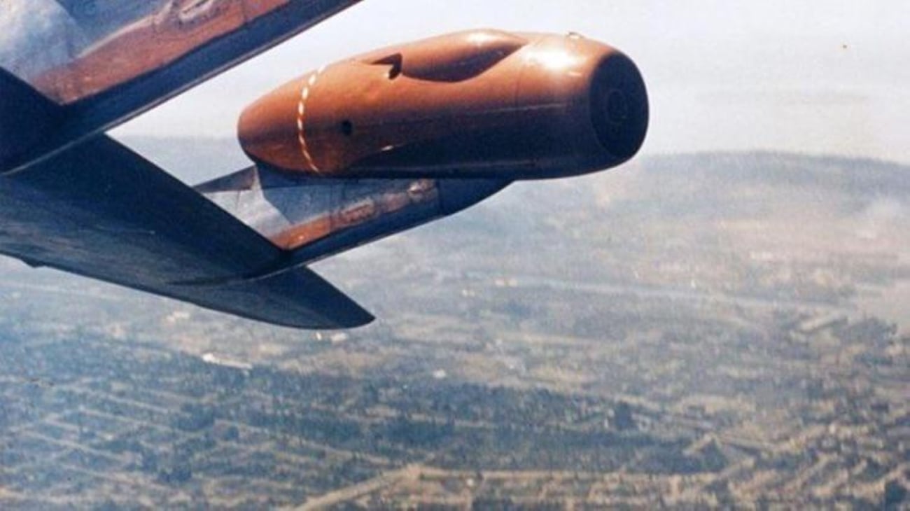 Aerial view of a landscape with a close-up of a jet engine mounted on an aircraft wing, flying above fields and scattered buildings, as seen through airplane windows.