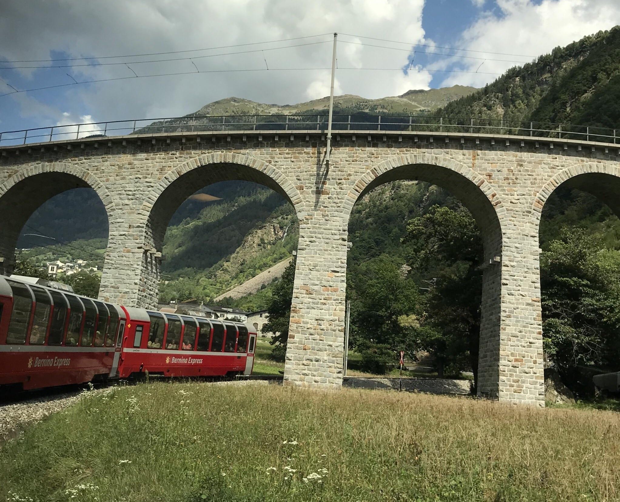 A red Bernina Express train curves beneath a stone viaduct with three arches, set against a backdrop of green hills, trees, and partly cloudy skies.