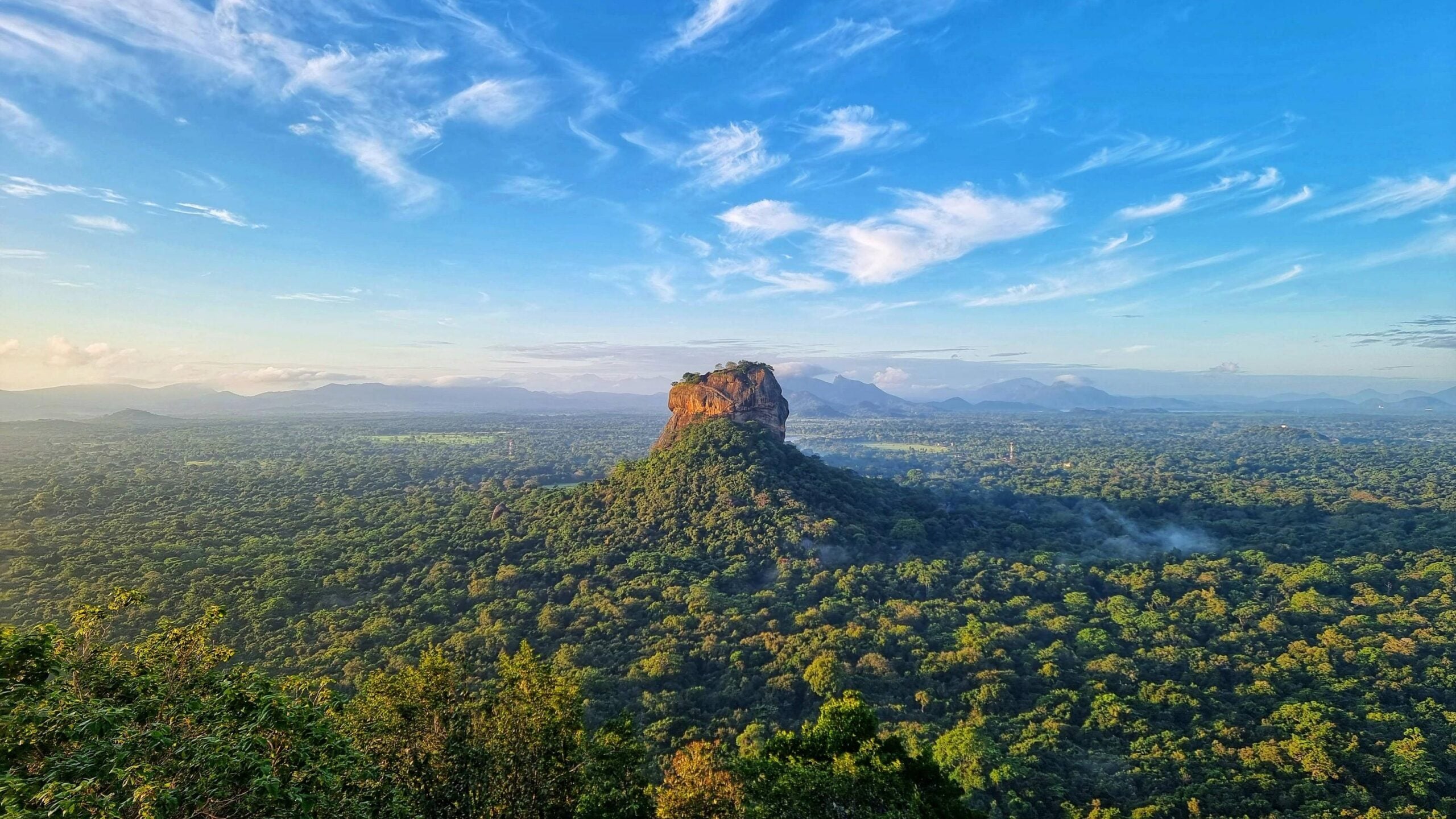 A large rock formation rises above a vast green forest under a bright blue sky with wispy clouds; distant mountains line the horizon, evoking the dramatic beauty often found in tropical locations.