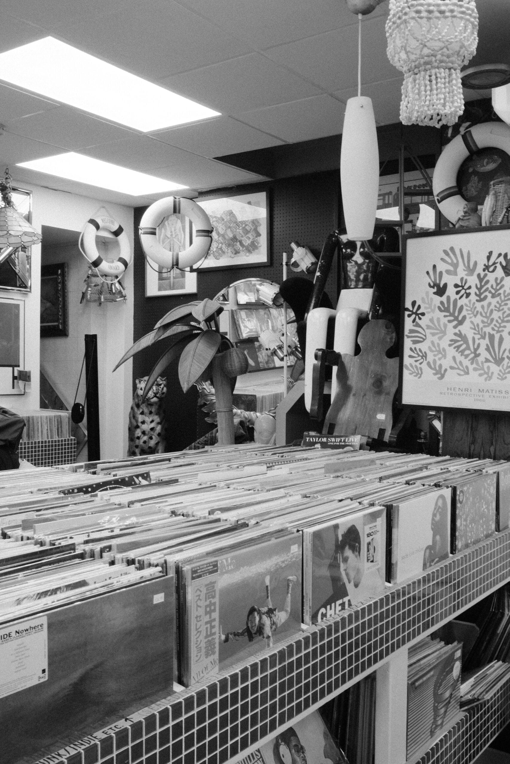 Black and white photo of a record store interior with rows of vinyl records, decorative plants, nautical life rings, and various hanging items, creating a cluttered and eclectic atmosphere.