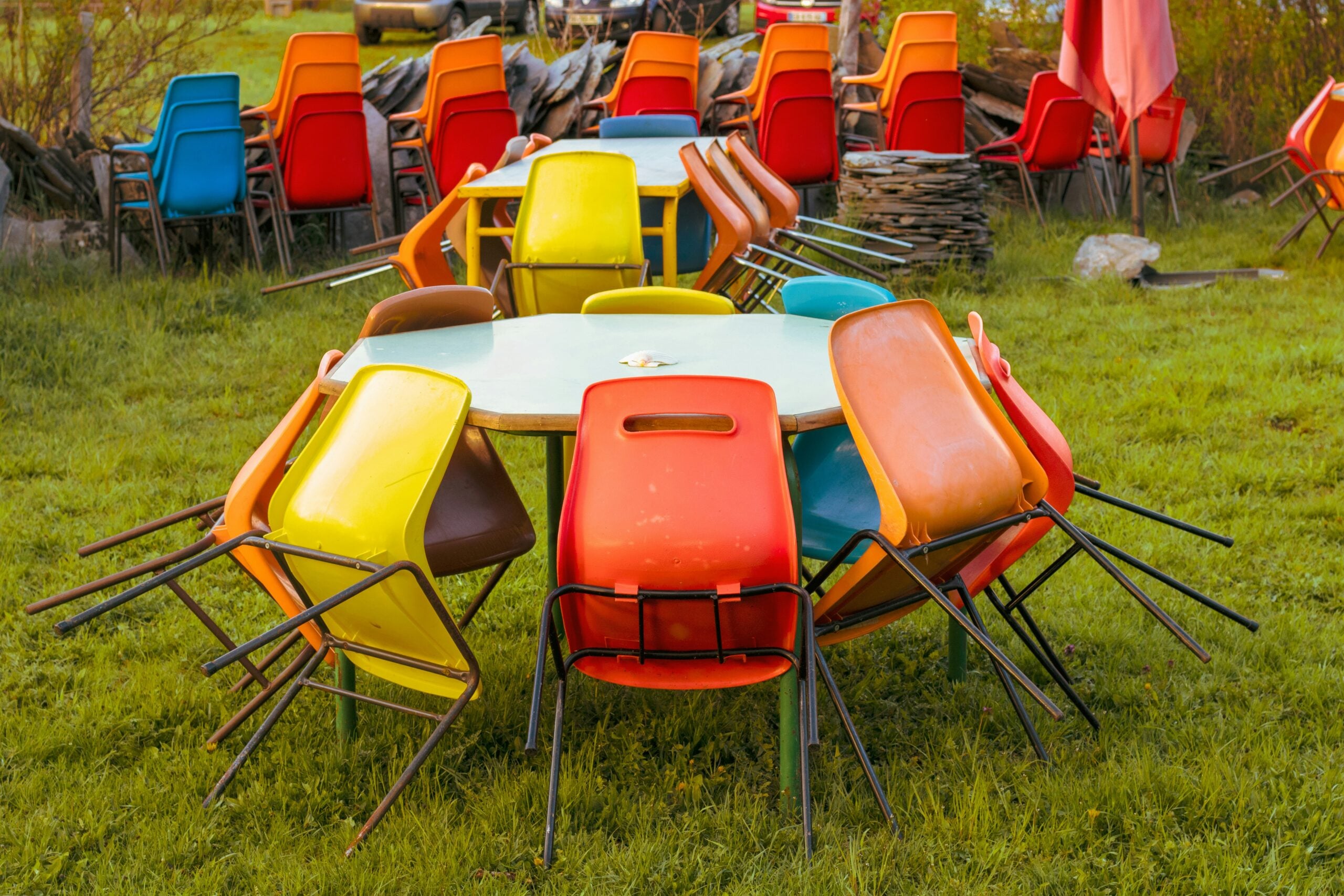 Colorful plastic chairs are stacked around round tables on a grassy lawn, with more chairs and tables grouped in the background, suggesting an outdoor event or gathering area.