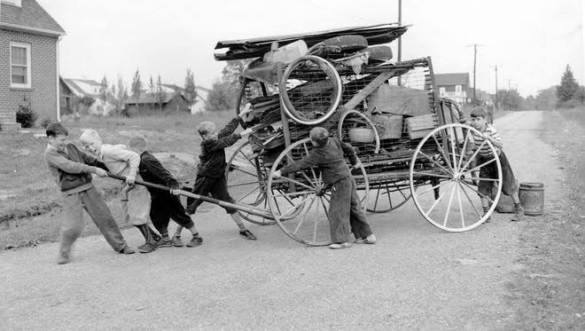 Six children push and pull a large, makeshift wooden cart overflowing with various objects and wheels along a quiet rural road.