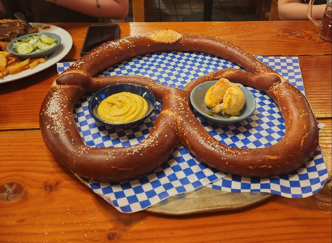 A giant soft pretzel on a blue checkered tray with two small bowls of mustard and cheese dipping sauce, placed on a wooden table.