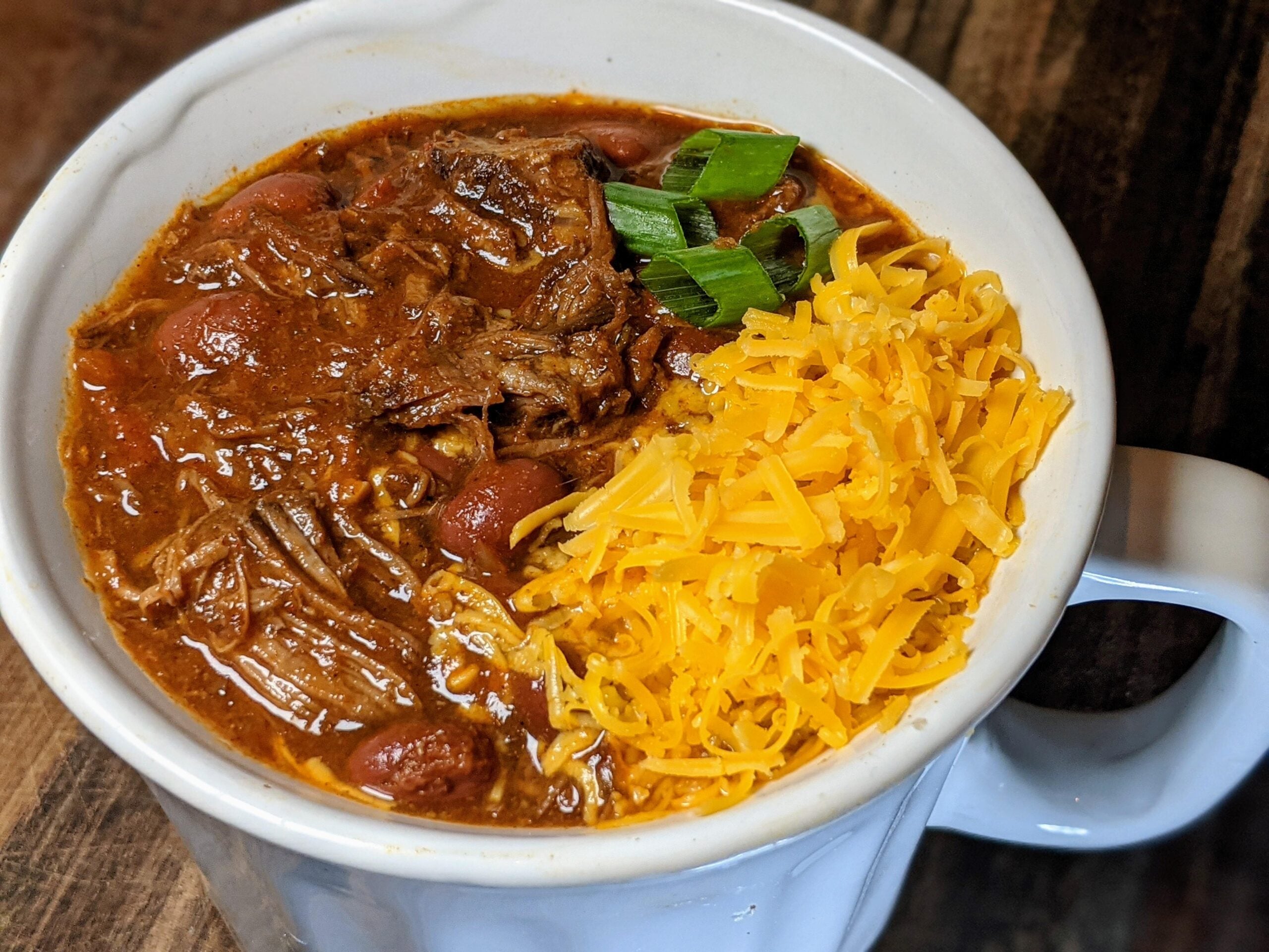 A white bowl filled with chili, topped with shredded cheddar cheese and chopped green onions, sits on a wooden surface.