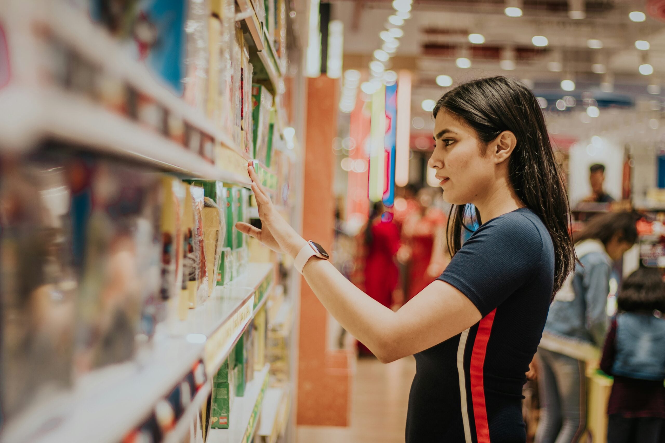 A woman with long dark hair browses items on a brightly lit store shelf, reaching out to touch a product. Other shoppers are visible in the background.