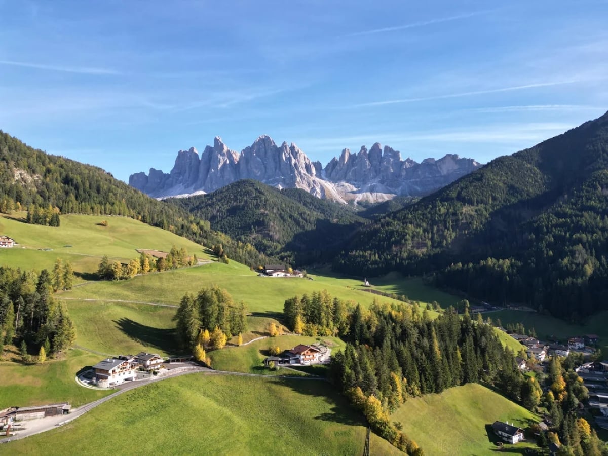 Rolling green hills with scattered houses, forested areas, and a dramatic rocky mountain range in the background under a clear blue sky.