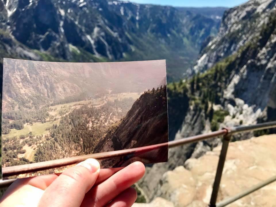A hand holds a photo of a valley landscape, aligning it with the same view in the background—showcasing how hikers have posted photos over time to capture changes in the scenery from an elevated viewpoint.