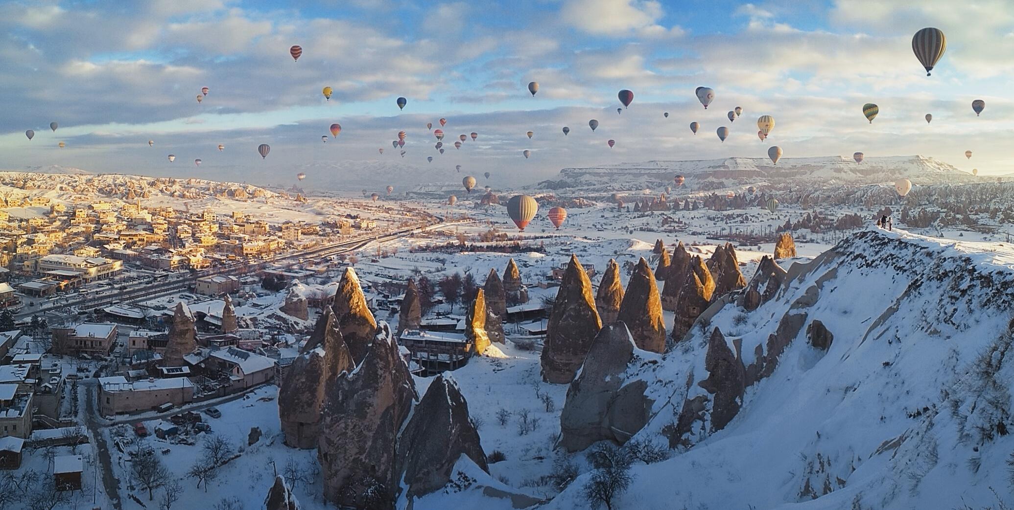 Colorful hot air balloons float over a snowy landscape with unusual rock formations and a town, under a partly cloudy sky in Cappadocia, Turkey.