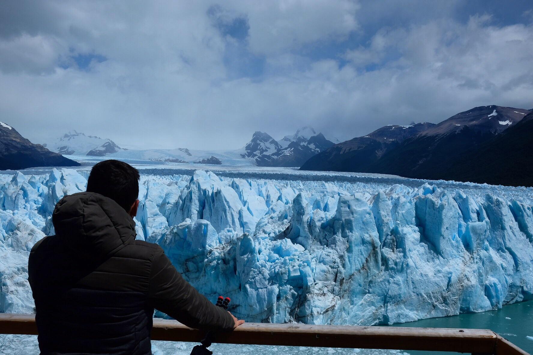 A person in a dark jacket stands at a wooden railing, looking out at a vast blue glacier with snowy mountains in the background under a partly cloudy sky.