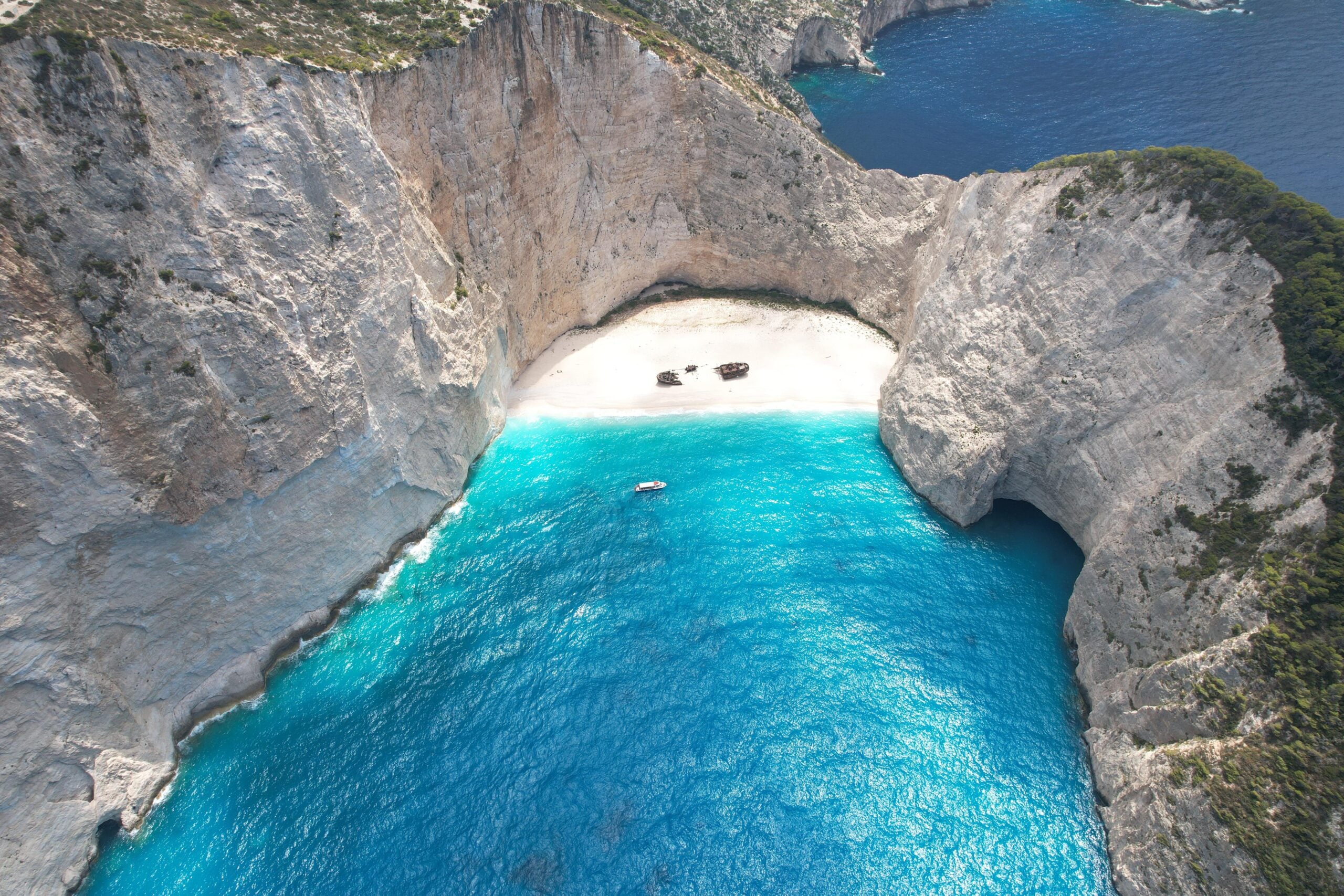 Aerial view of a small sandy beach surrounded by high cliffs, with turquoise blue water and a shipwreck on the sand—one of the region’s most incredible beaches. A boat floats nearby in the sea.