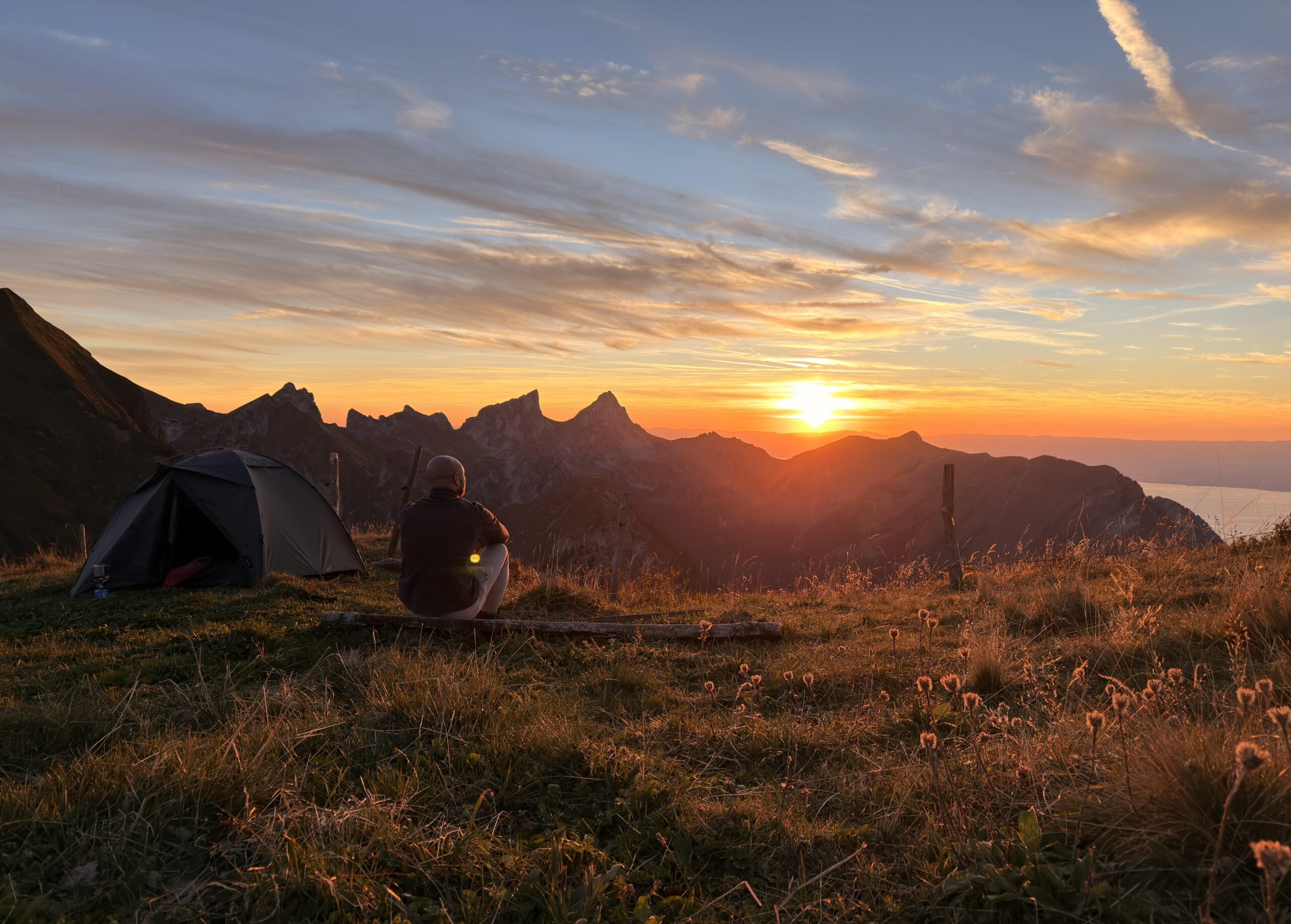 A person sits beside a tent on a grassy hill, watching the sun set behind rugged mountains. The sky is filled with orange and blue hues, and wildflowers are softly lit in the foreground.