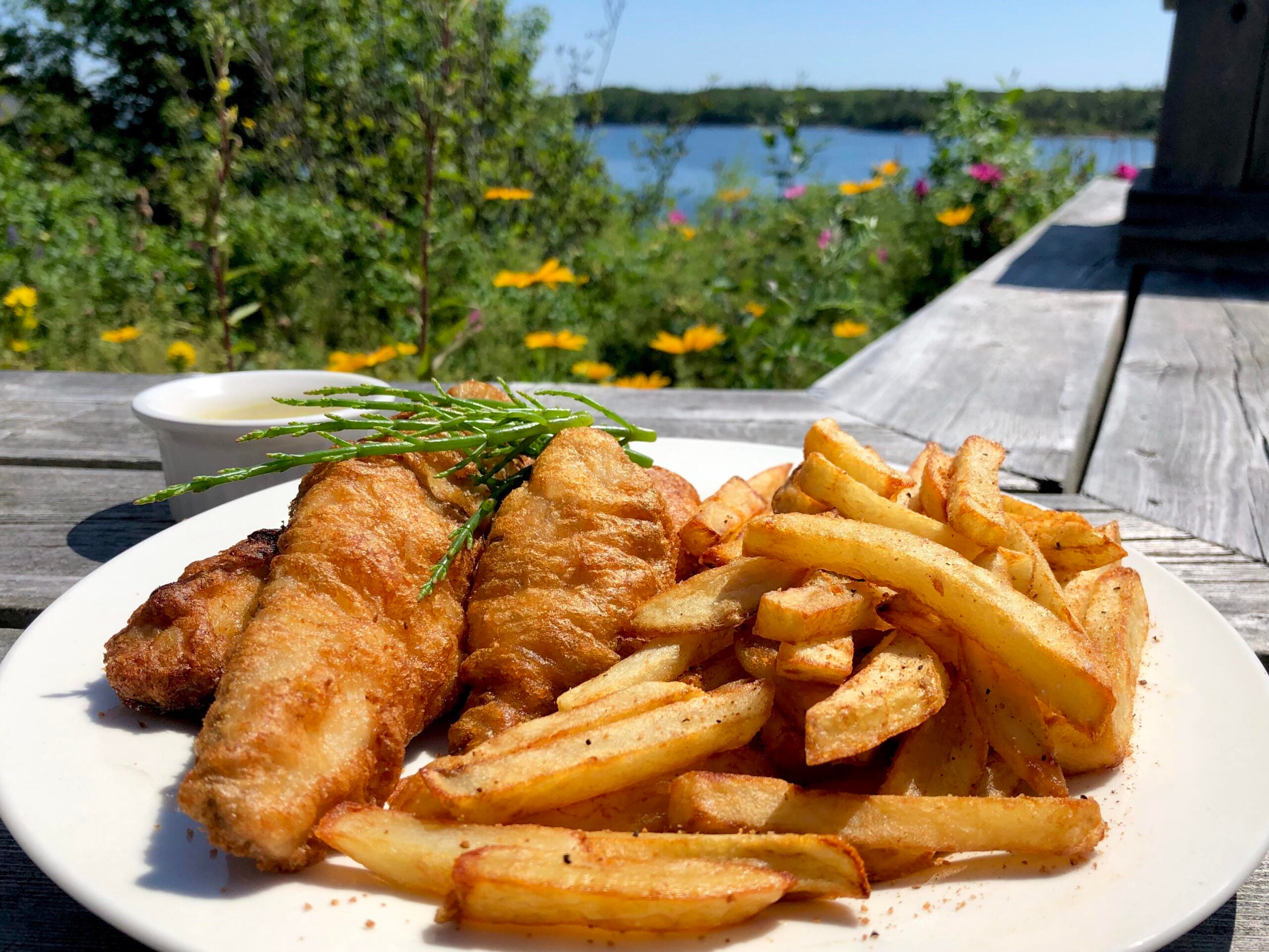 A plate of golden fried fish and seasoned fries sits on a wooden table outdoors, with wildflowers and a blue lake visible in the sunny background.