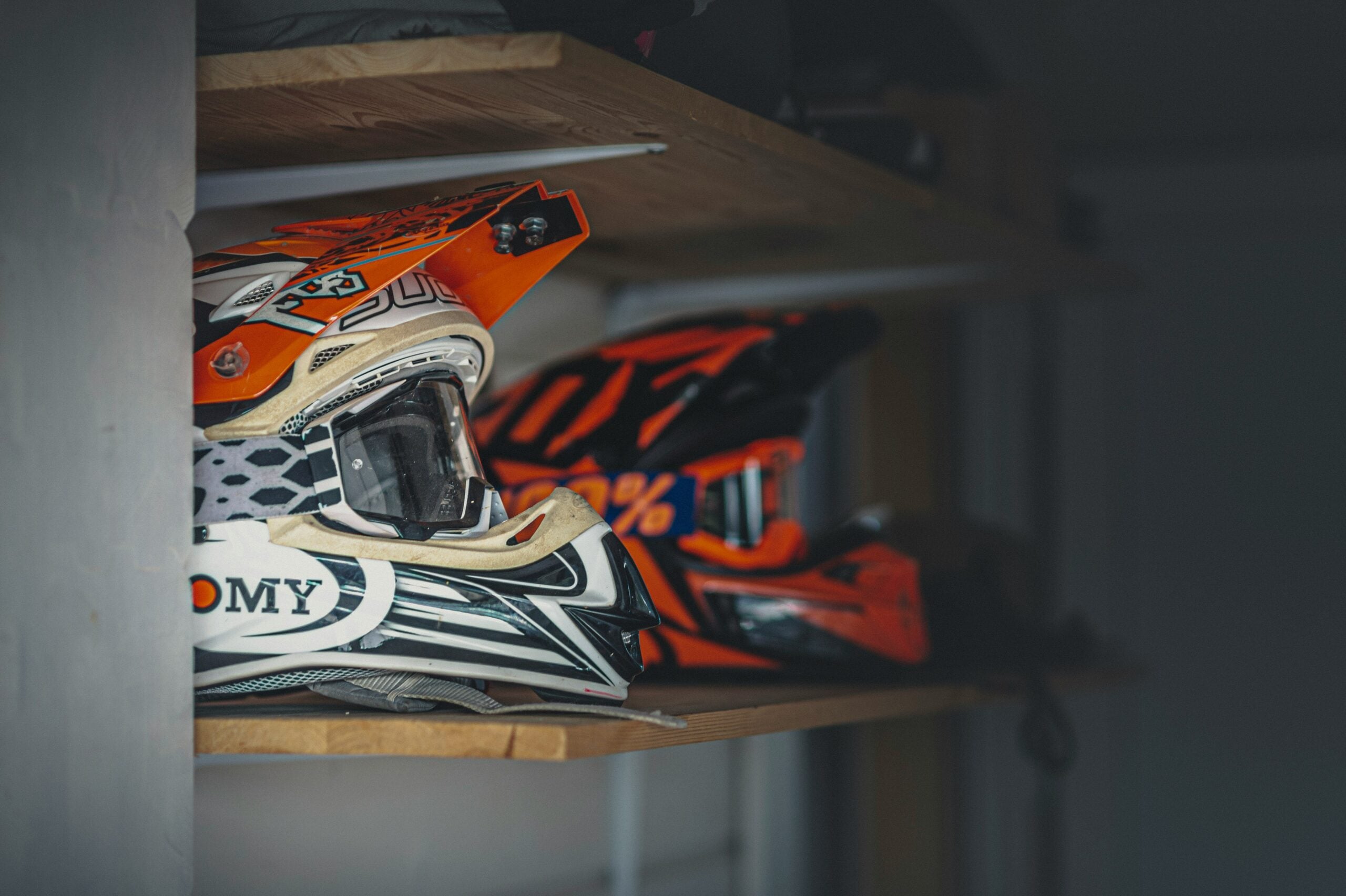 Two brightly colored motocross helmets, mostly orange and white, sit on a wooden shelf in a dimly lit room. The helmets feature visors and goggles, and the focus is on the helmet in the foreground.