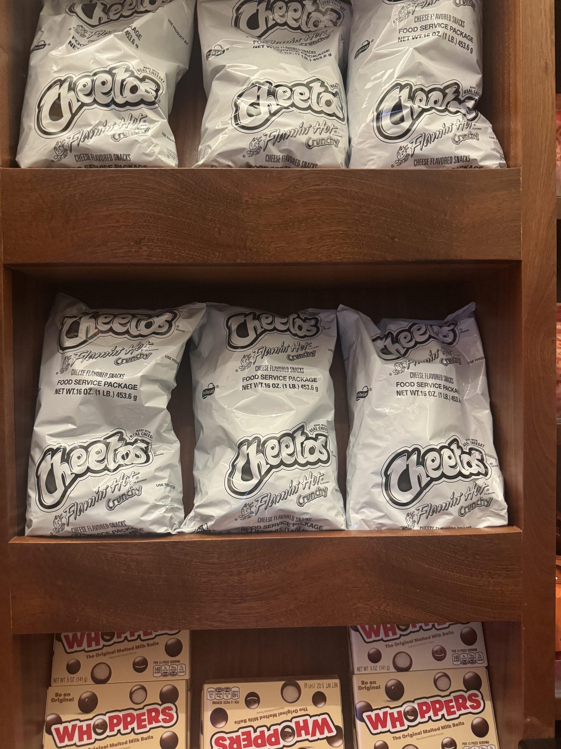 Bags of Flamin’ Hot Cheetos in monochrome packaging sit on wooden shelves above boxes of Whoppers malted milk balls in a store display.