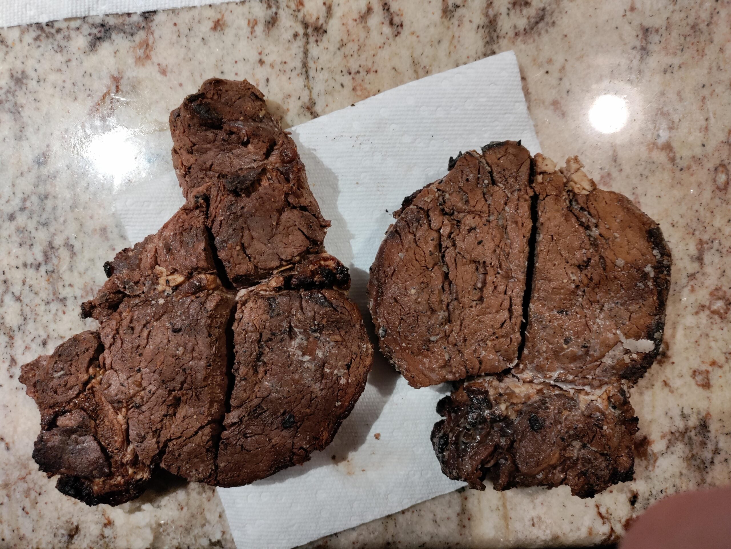 Two large, dark brown, cracked cookies rest on a white paper towel atop a marble countertop. The cookies have a rough texture and uneven shapes, with visible pieces baked into the dough.