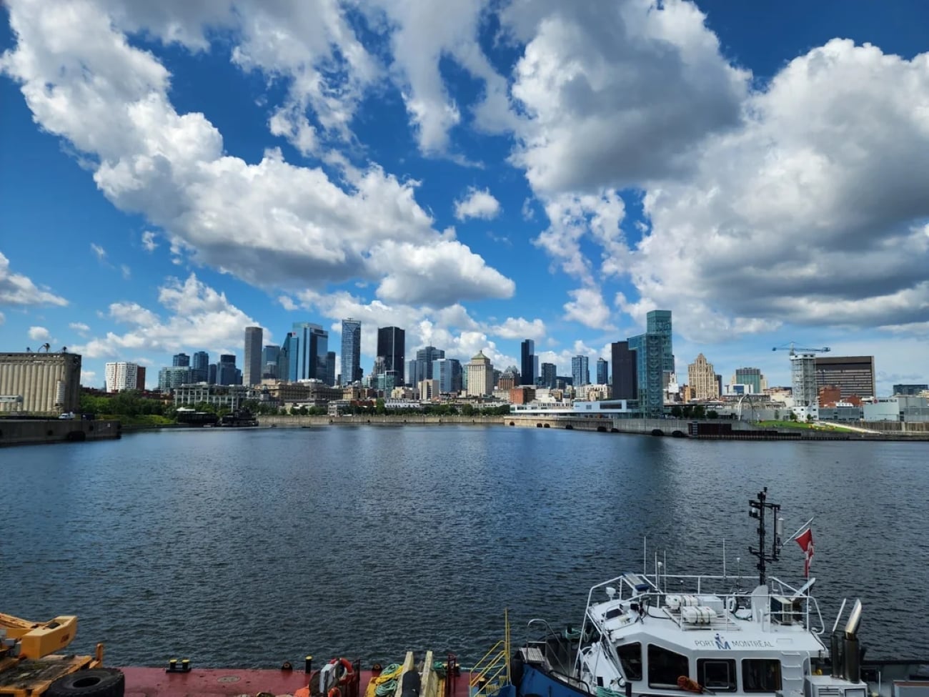 A city skyline with tall buildings under a blue sky with white clouds, viewed across a river with boats docked in the foreground.