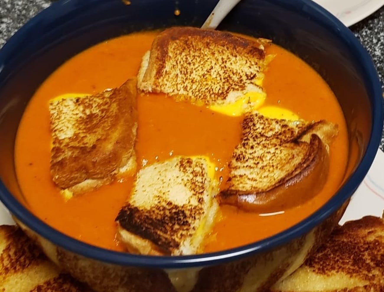 A bowl of tomato soup with pieces of grilled cheese sandwiches floating on top, featuring classic grilled cheese sandwiches and a spoon resting inside the bowl.