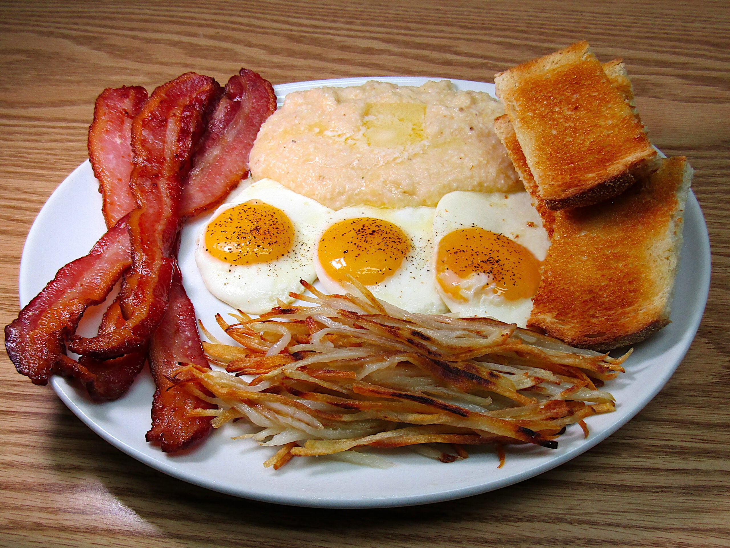 A plate with crispy bacon strips, three sunny-side-up eggs, buttered grits, golden toast slices, and a serving of hash browns on a wooden table.