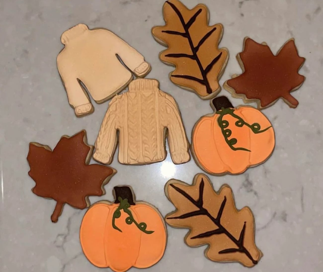 Assorted decorated cookies shaped like sweaters, pumpkins, and leaves are arranged on a light gray marble surface, representing an autumn theme.