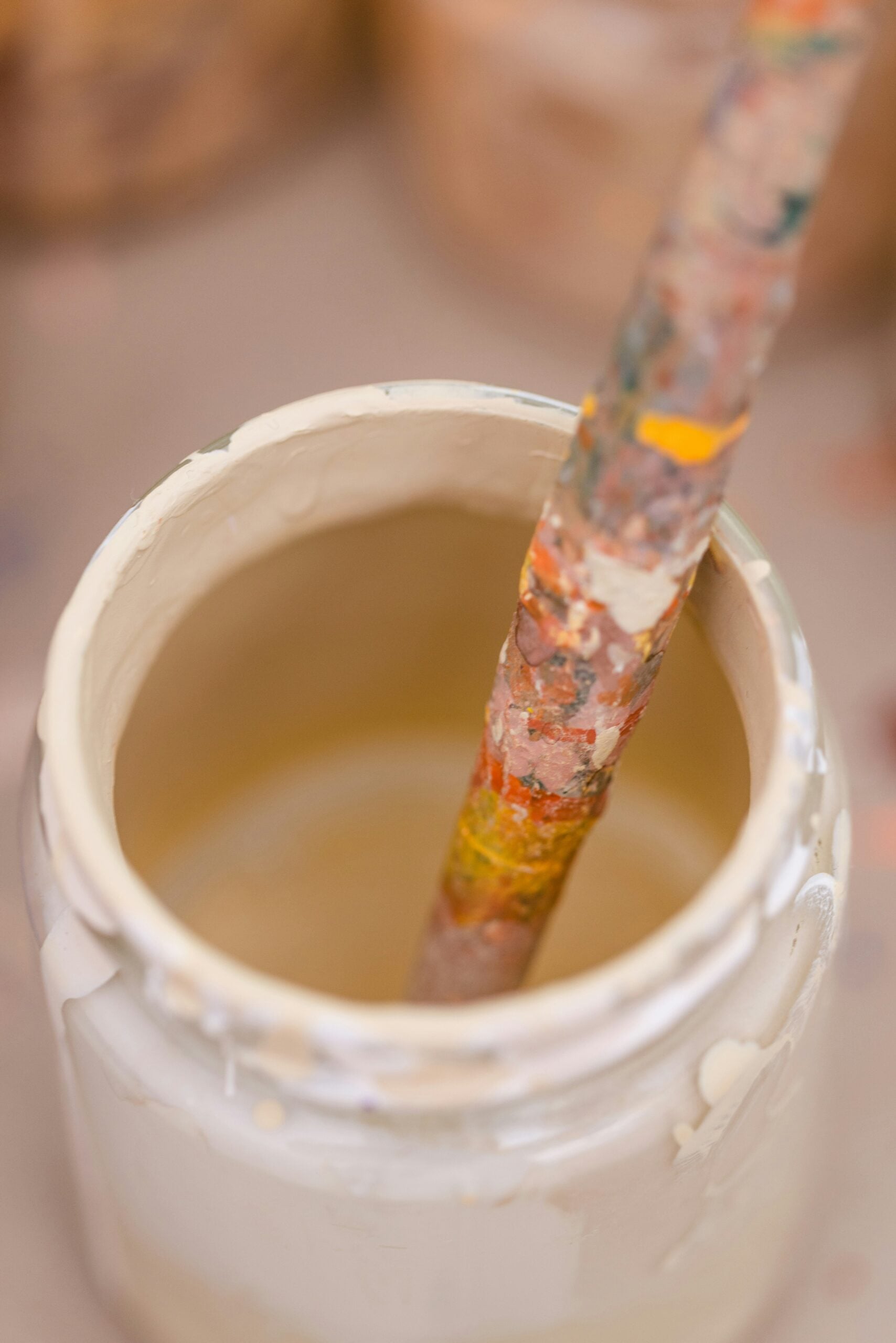 A close-up of a paint-splattered brush resting inside a white paint jar, with blurred containers in the background.