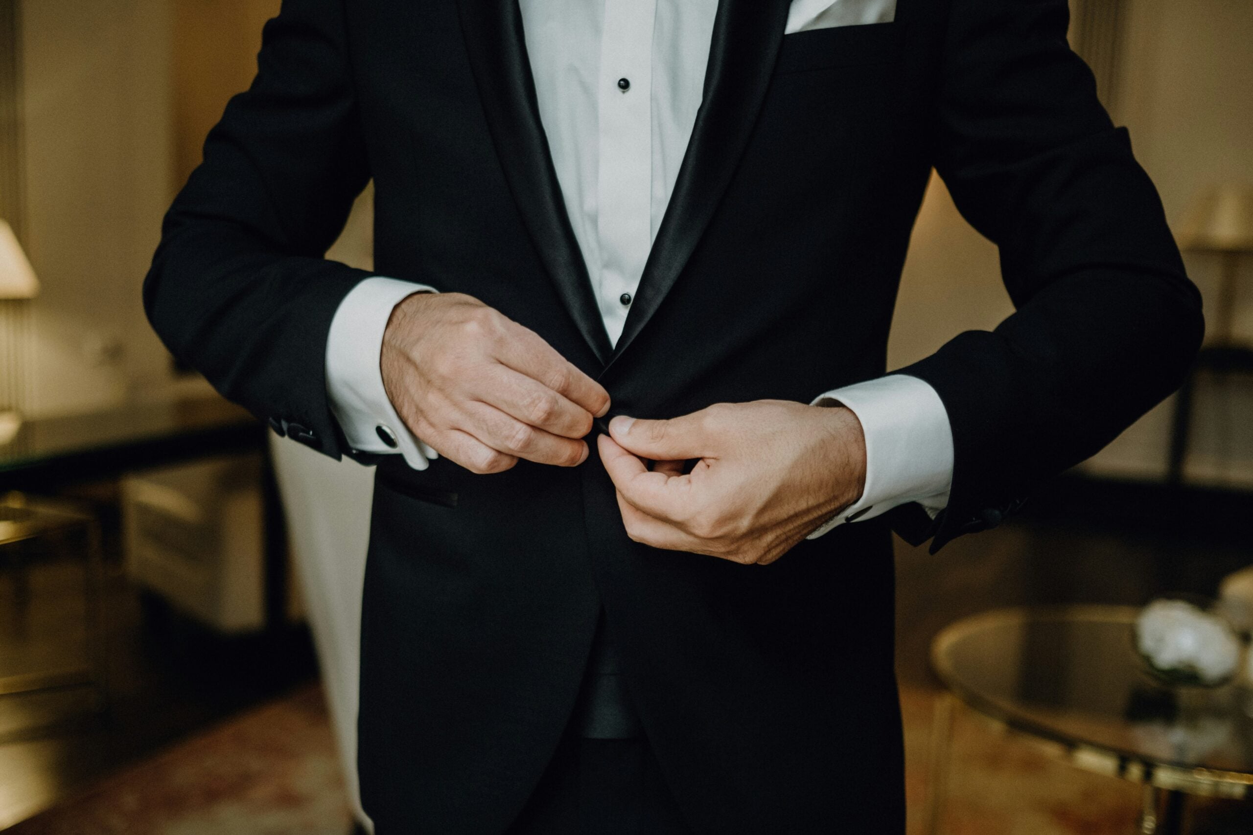 A person in a black tuxedo is buttoning their jacket. The image focuses on their hands and torso, with a blurred background of a room with furniture.