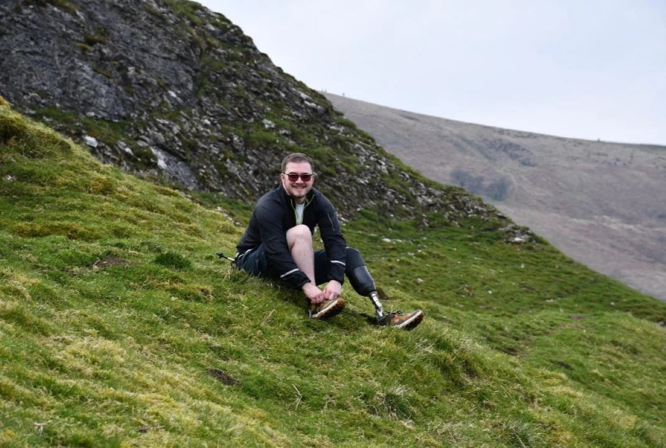 A man with prosthetic legs sits on a grassy hillside, smiling and adjusting his shoe. Rocky terrain and a sloping hill are visible in the background under a cloudy sky, creating the perfect scene for photos often posted by hikers who embrace adventure regardless of obstacles.