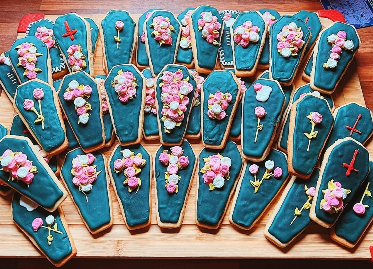 A group of coffin-shaped cookies decorated with dark icing, colorful flower designs, and some with red crosses, arranged on a wooden surface.