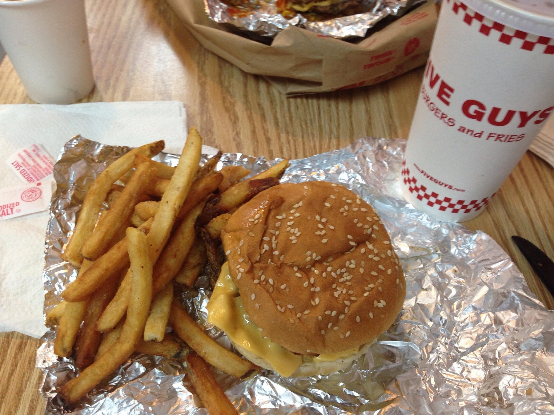 A cheeseburger with a sesame seed bun and a serving of French fries on foil, next to a large Five Guys drink cup, on a wooden table.