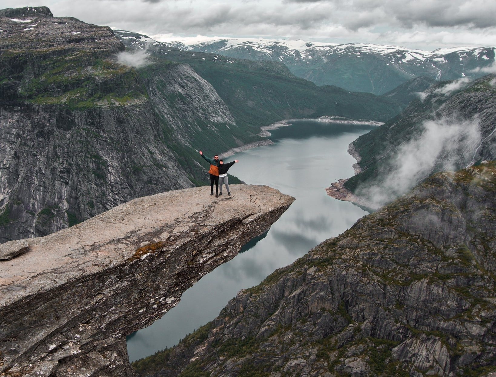 Two people stand with arms raised on a dramatic rock ledge overlooking a deep fjord and steep green mountains under a cloudy sky.