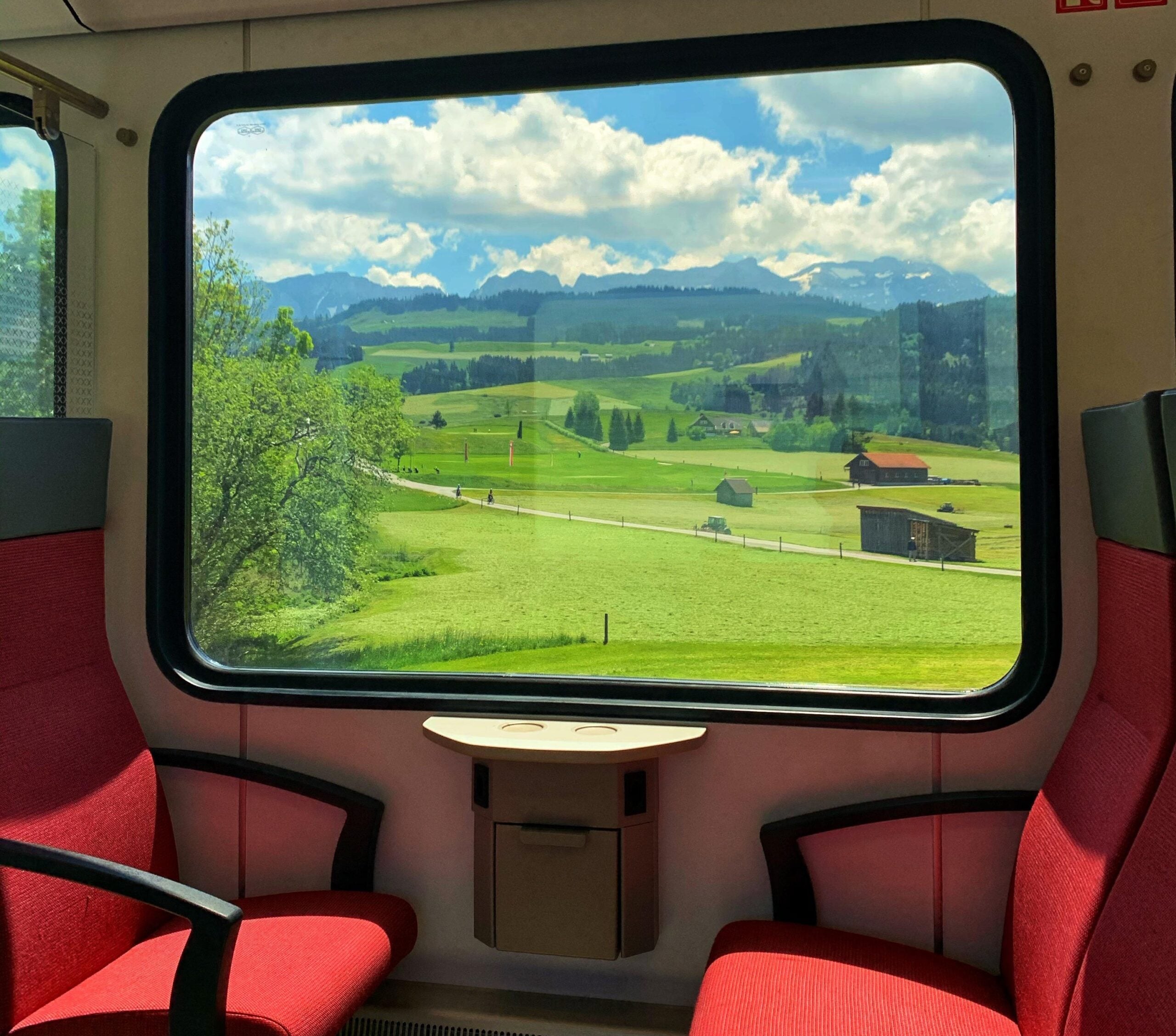 A scenic view of green fields, scattered houses, and mountains is seen through a large train window. Two red seats and a small table are inside the train.
