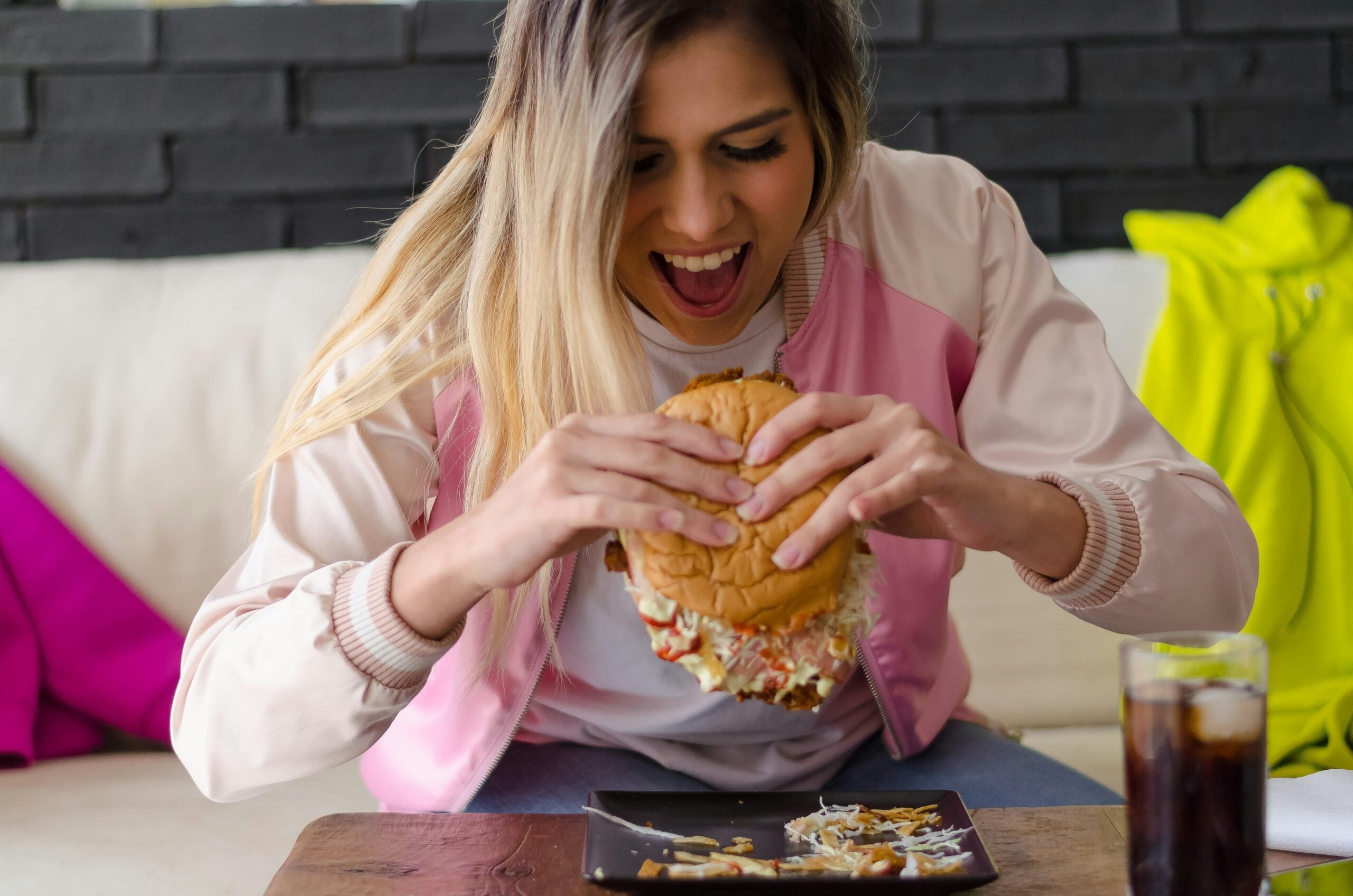 A woman in a pink jacket excitedly prepares to eat a large, messy hamburger, sitting on a couch with a plate and a glass of soda in front of her.