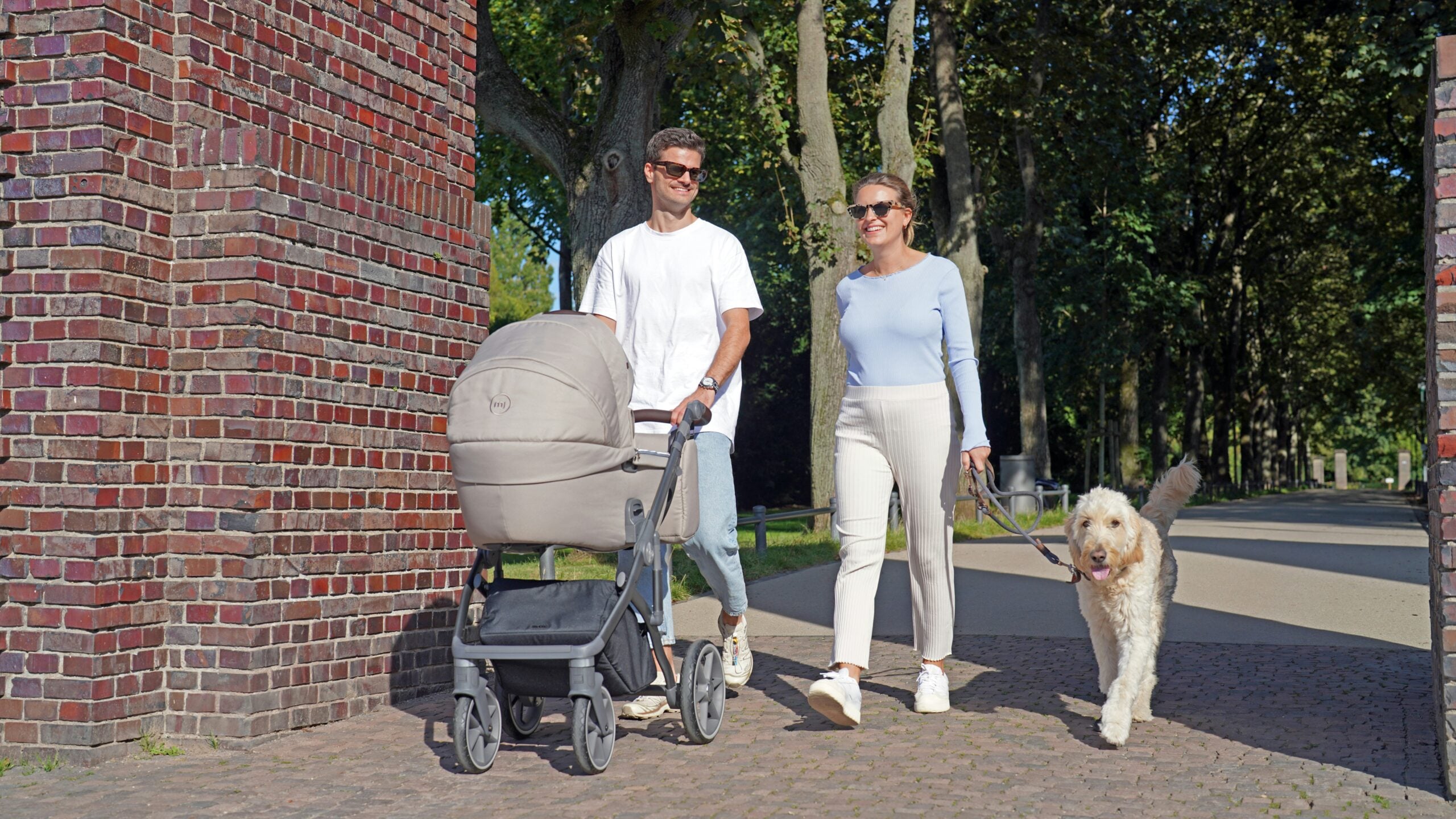 A man and woman walk outside; the man pushes a stroller and the woman holds a dog’s leash. They are smiling, and there are trees and brick columns in the background.