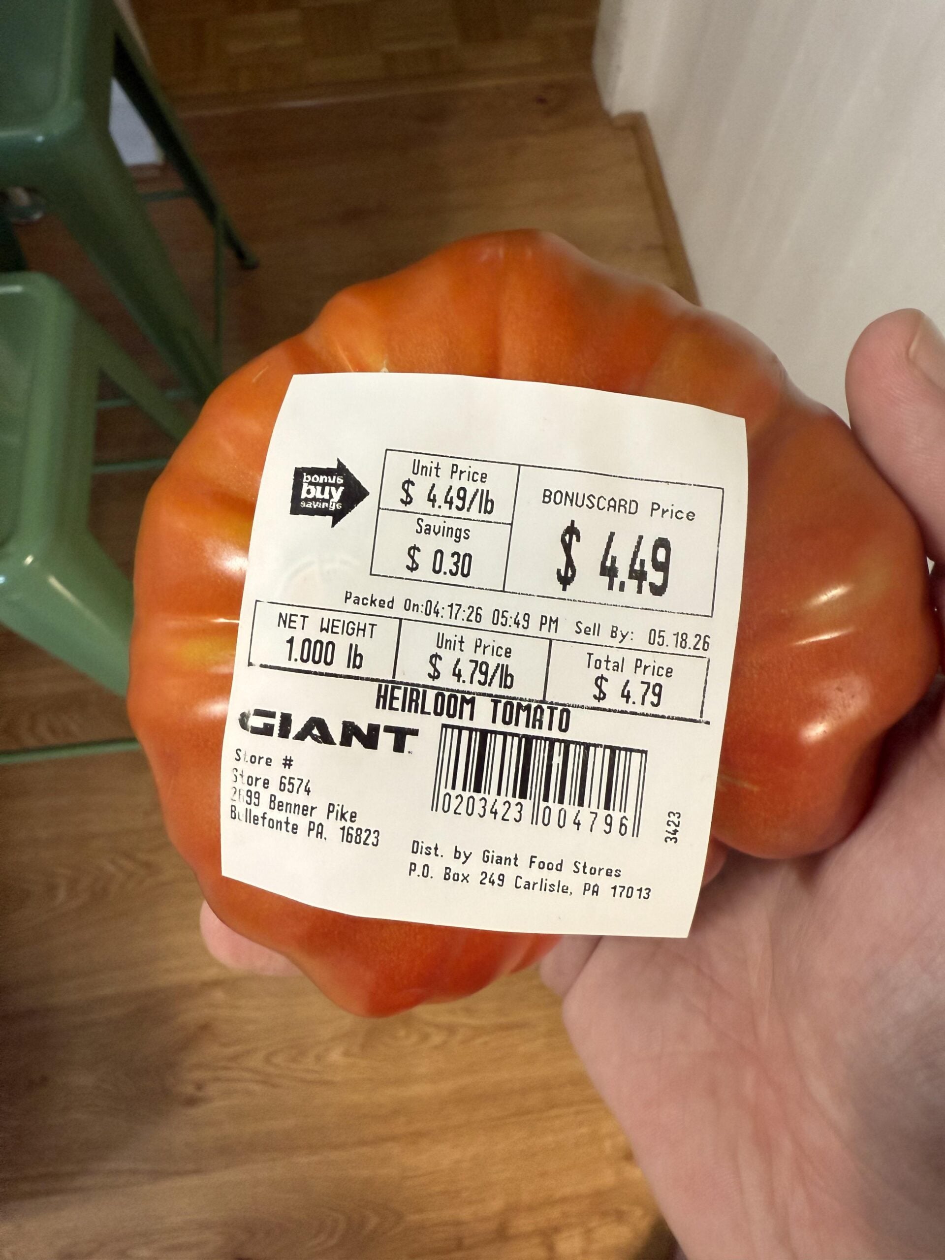 A hand holds a large heirloom tomato with a white price label showing $4.49 per pound and total price $4.49. Green chairs and a wooden floor appear in the background.