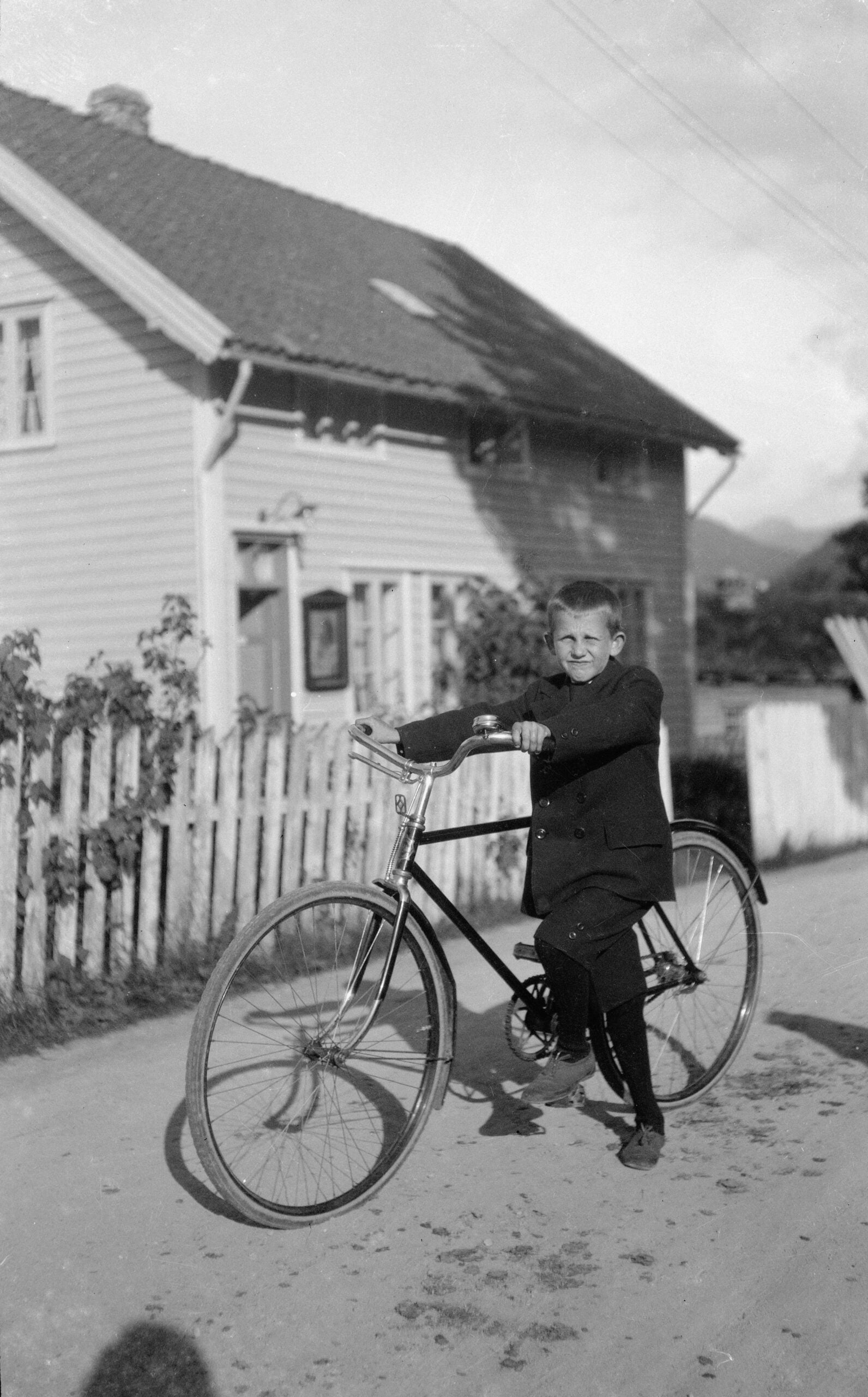 A young boy in a suit stands with a bicycle on a dirt road in front of a wooden house and picket fence, smiling at the camera in bright daylight.
